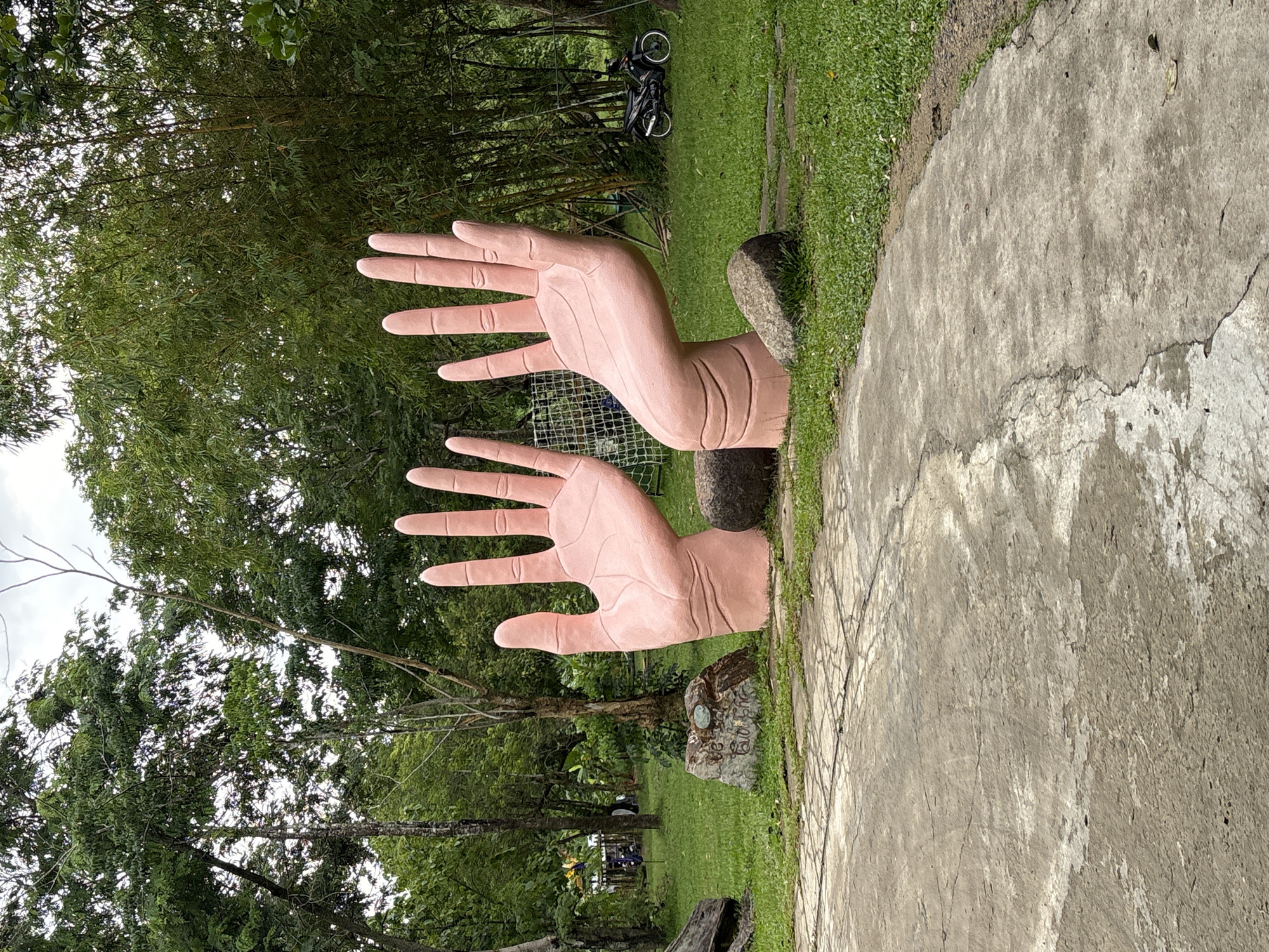 Two large, pink, sculptural hands rise from a grassy area within a park. The left hand is raised and open, with fingers splayed, while the right hand is bent at the wrist, with fingers also spread. Both sculptures appear to be made of a smooth, solid material. They are positioned on a concrete path that curves through the park. In the background, lush green trees and bamboo stalks dominate the landscape. A black motorcycle is parked on the grass to the right. A few large rocks are scattered around the base of the hand sculptures. The sky is overcast. The location is identified as Khanh Vinh, Vietnam.