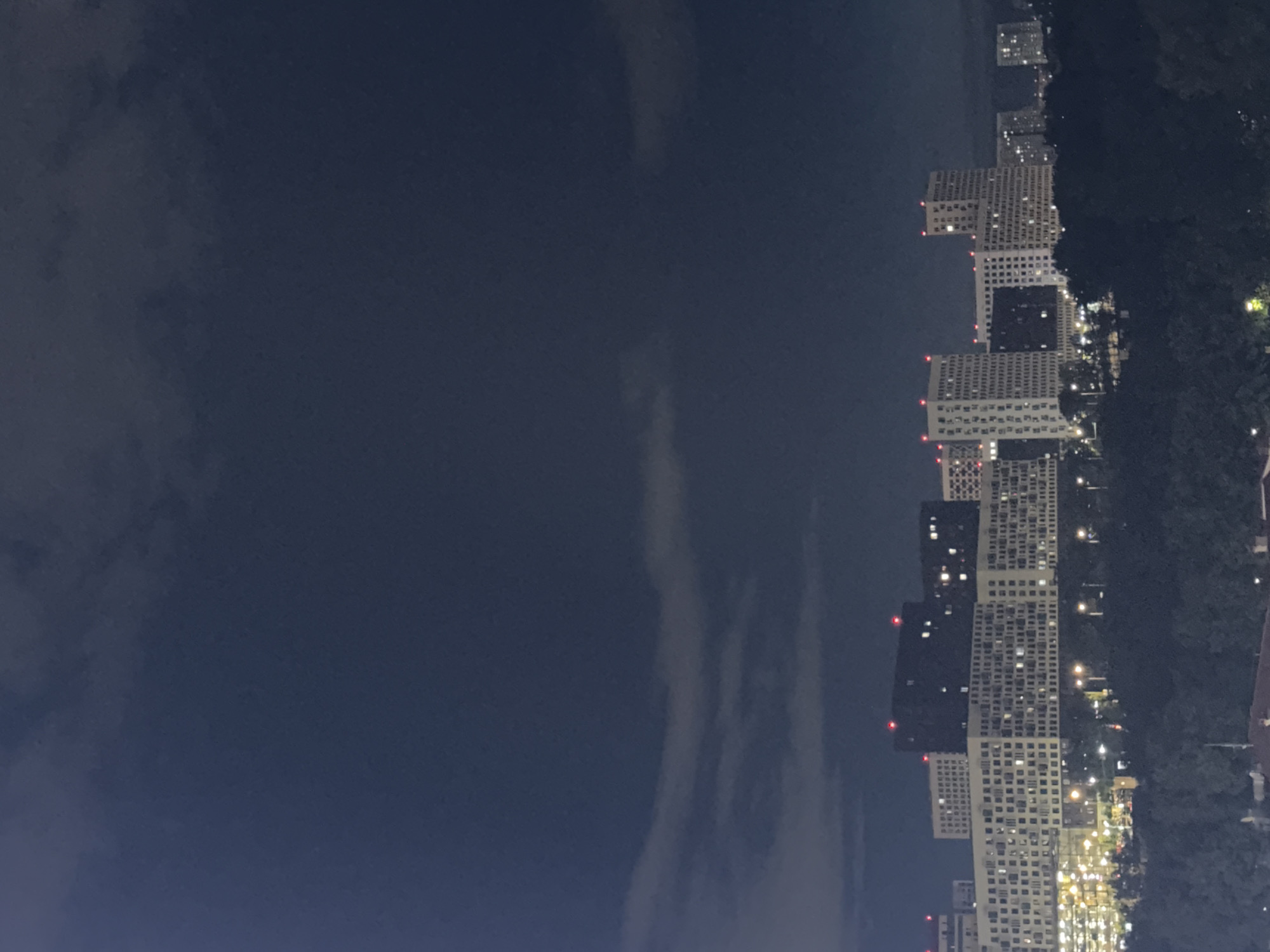The image shows an urban landscape at night, likely taken in Khimki, Russia. The right side of the frame is dominated by a row of high-rise apartment buildings, illuminated with many lights suggesting residential activity. Some buildings are white with a grid-like pattern of windows, while others are darker. Red lights, possibly obstruction lights for aviation safety, are visible atop some buildings. Adjacent to the buildings on the right is a line of trees. The left side of the image shows a night sky with some thin cloud formations. The overall impression is an urban night scene.