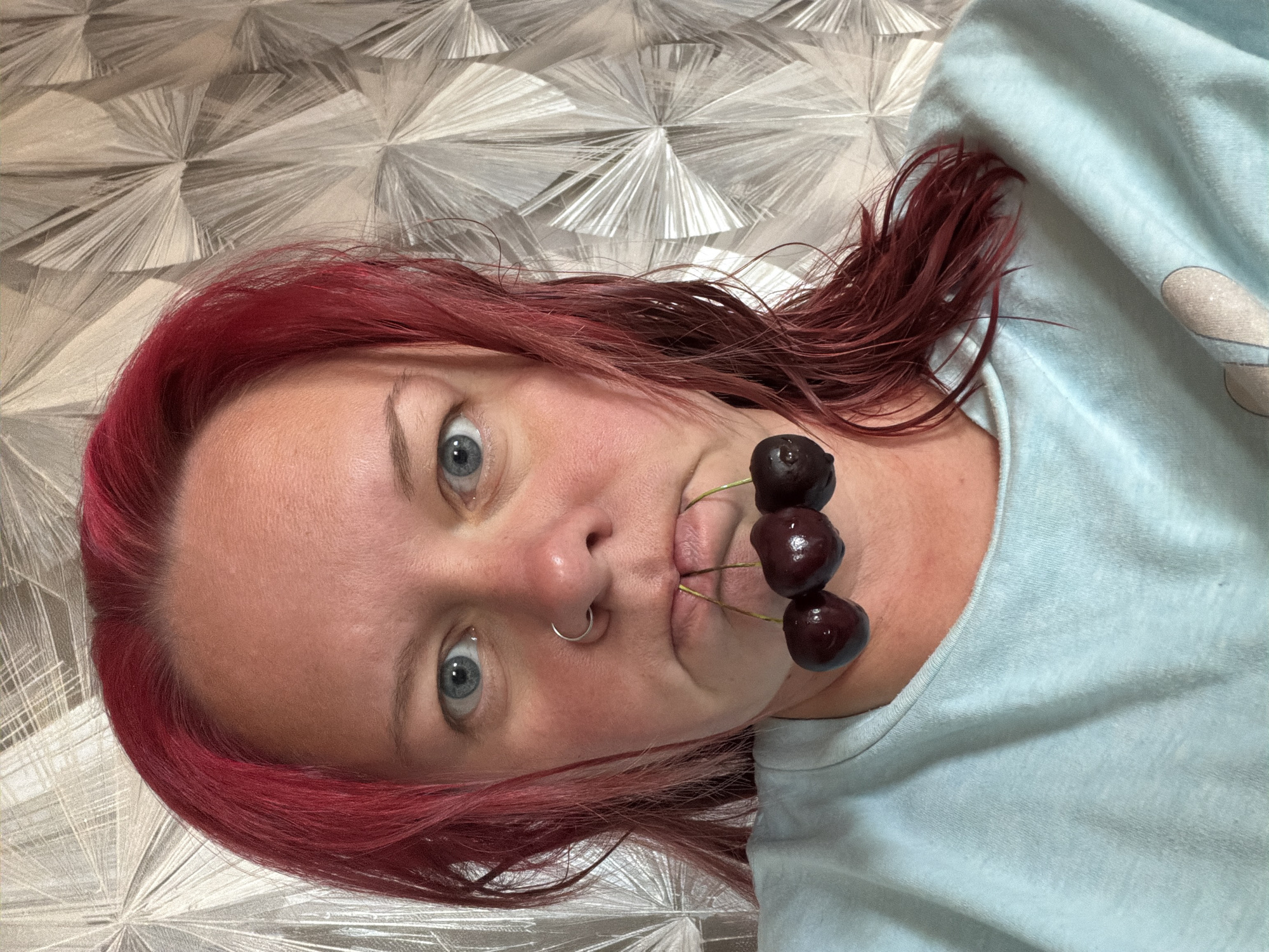 A medium close-up shot shows a woman with bright red hair and blue eyes. She is wearing a light blue crewneck shirt and has a silver hoop ring in her nose. Three dark red cherries with stems are held between her lips. The background is a patterned wallpaper in shades of gray and white. The image is a selfie, suggesting the woman is in an indoor setting, likely a home, and it is not possible to determine the specific location in Chaikovsky, Russia from the visual information provided.