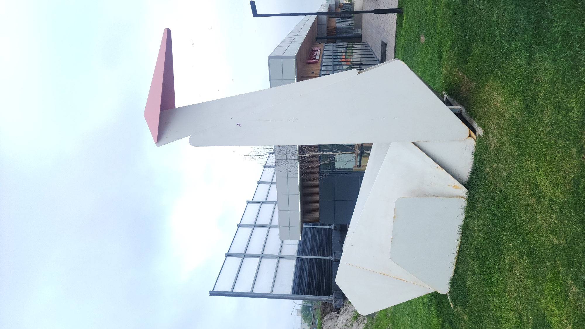 The image shows a large, modern, abstract sculpture made of white metal plates with a pink triangular accent piece. The sculpture is situated on a patch of green grass, likely in a public space. In the background, there are modern buildings with glass windows and metal framework. The sky is overcast. The scene is likely in an urban environment, potentially Moscow, Russia. The composition of the image suggests the photographer's intention was to capture the sculpture against the backdrop of the modern architecture and overcast sky.
