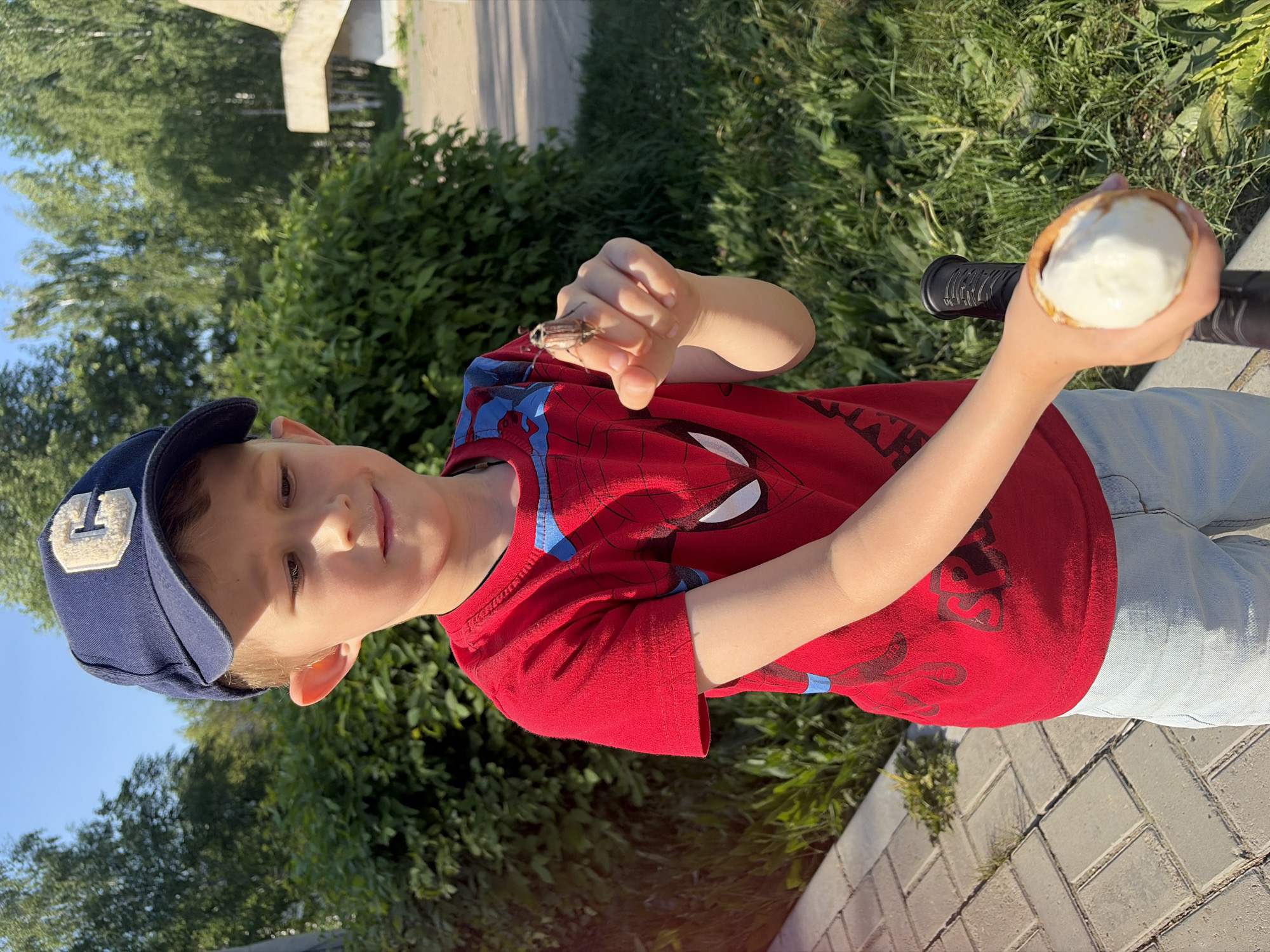 A young boy in a red Spiderman shirt and a blue cap holds an insect on his hand and an ice cream cone in the other. He stands on a brick pathway, with green bushes in the background. The location is outdoors, likely in a park or residential area, possibly in the city of Chaikovsky, Russia.