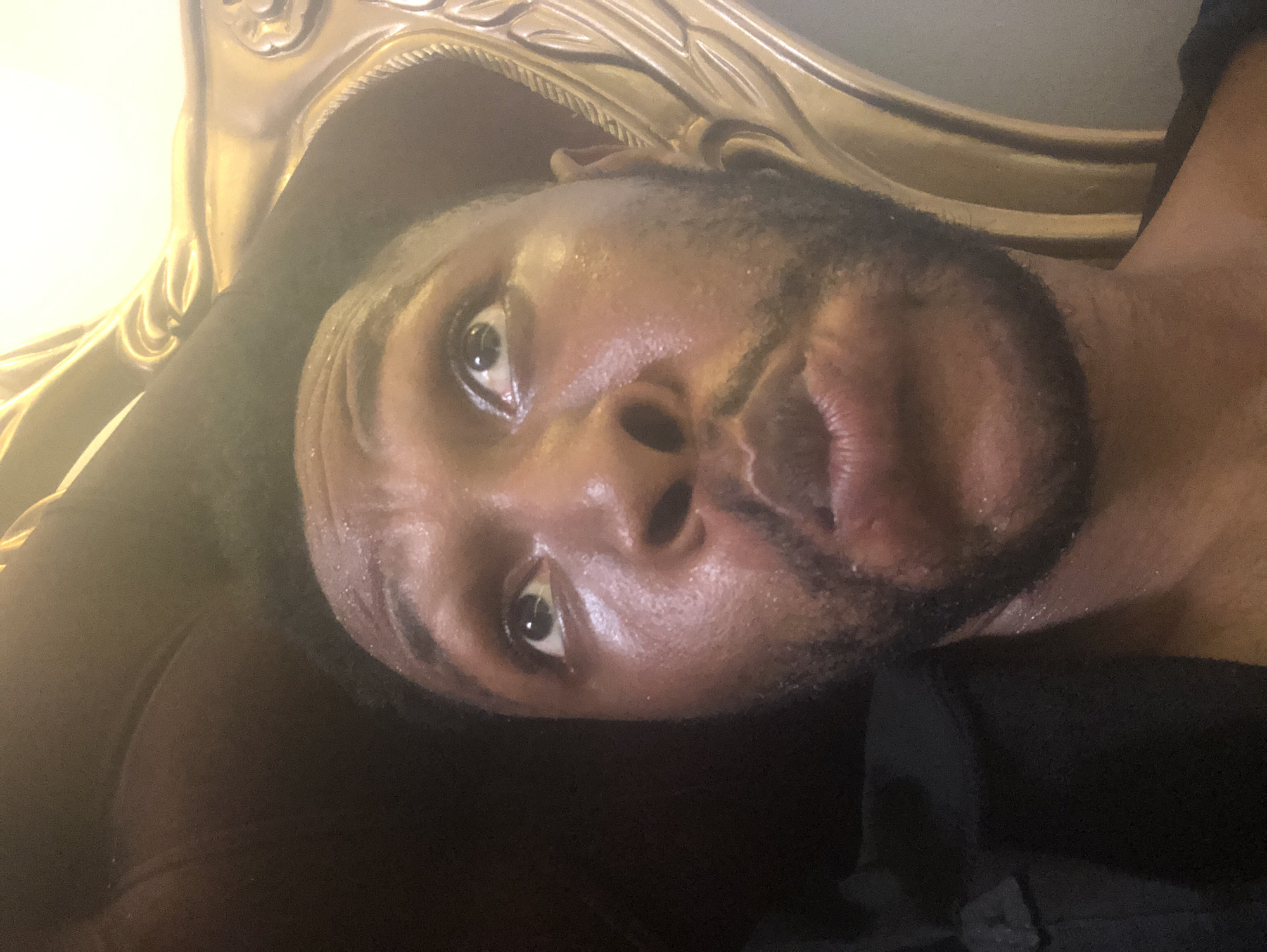 An upward-angled, close-up shot captures a dark-skinned male subject reclining or lying down, looking directly towards the camera or slightly past it. The image is rotated approximately 90 degrees clockwise, placing the subject's head on the left side of the frame.
The man has a short beard and moustache. His eyes are open and appear to be focused. His skin, particularly on his forehead and around his nose and mouth, shows a noticeable sheen, possibly from sweat or natural oils, reflecting the warm light. His lips are slightly parted. He is wearing a dark-colored garment, likely a shirt or t-shirt, which is visible across his shoulder and chest on the right side of the frame.
Behind the subject's head and upper body, an ornate, gold-colored piece of furniture is visible, likely a chair, sofa, or headboard, featuring intricate carved details and a rich, shiny texture. The background suggests an indoor setting. The lighting is warm, casting a gentle glow that highlights the contours of the subject's face and the details of the furniture. There is no visible text in the image.