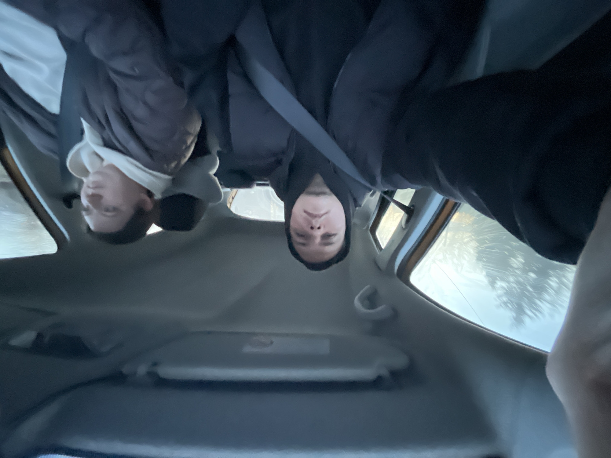 The image depicts the interior of a car, with the photographer positioned upside-down relative to the car's orientation. Two people are visible: one wearing a dark jacket and a seatbelt, and another to their left wearing a lighter jacket with a hooded sweatshirt underneath. The car's sun visor is visible in the foreground. Outside the car windows, blurry shapes suggestive of trees or foliage can be seen. Location in Yasenevo, Russia cannot be confirmed from this image.