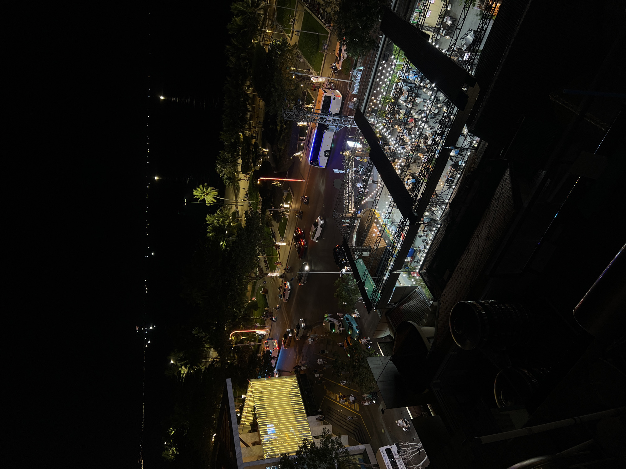 This is a nighttime, high-angle aerial view of a street scene in Nha Trang, Vietnam, likely along the coastal promenade. The image captures a vibrant urban environment with a mixture of buildings, vehicles, and pedestrians.
In the foreground, the rooftops of several structures are visible, some appearing to be commercial establishments with covered outdoor seating areas illuminated by strings of lights. These areas are populated with many individuals seated at tables, suggesting a lively dining or social scene.
Further back, a street is illuminated by various lights, including streetlights, building lights, and vehicle headlights. Several cars and motorcycles are present, indicating traffic flow. A prominent building on the left is adorned with bright yellow vertical strip lights, creating a striking visual element.
Beyond the street, a dark expanse of water, likely the sea, stretches to the horizon. The lights of distant structures and possibly boats are visible on the water. A row of palm trees lines the area between the street and the sea, characteristic of a tropical coastal city. The overall scene depicts a bustling urban nightscape with elements of commerce, transportation, and leisure.