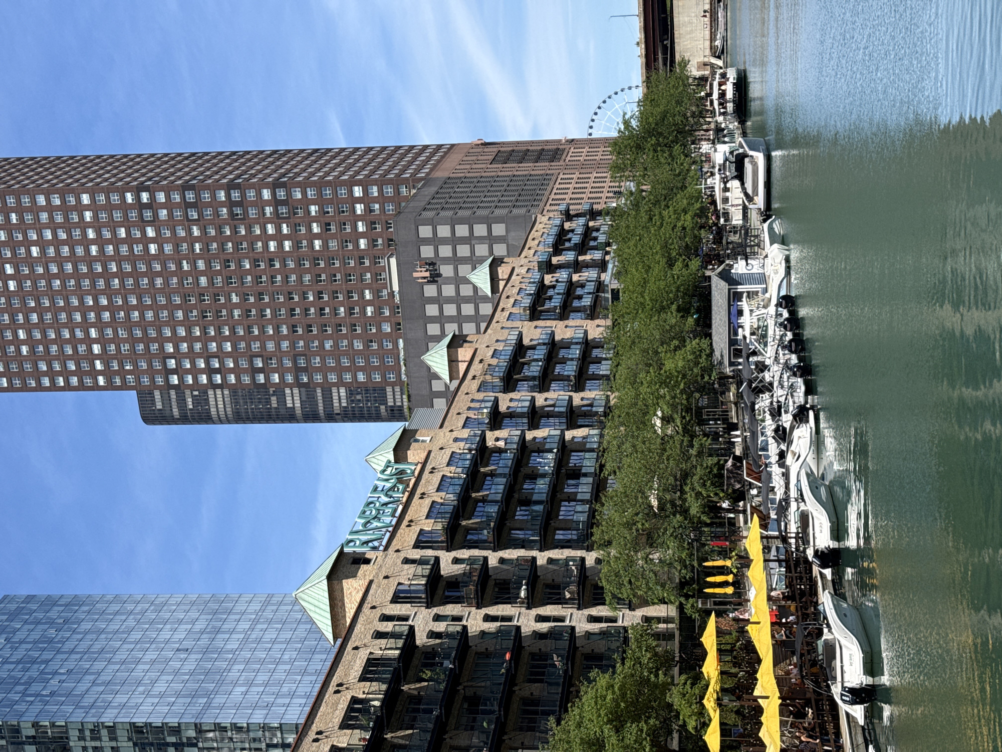 This image depicts a scenic view of the Chicago Riverwalk in Chicago, Illinois, USA. The foreground features several white motorboats docked along the river, their reflections shimmering on the turquoise water. To the left, a row of yellow umbrellas shades outdoor seating areas, indicating a vibrant dining and leisure scene. Behind the umbrellas, a multi-story brick building with many balconies and dark-paned windows lines the river. Above this, a contemporary glass skyscraper and a taller, brown brick skyscraper dominate the skyline, showcasing Chicago's iconic architecture. In the distance, a Ferris wheel can be seen, a landmark of the city. The sky is a clear blue with scattered white clouds, suggesting a bright, sunny day.