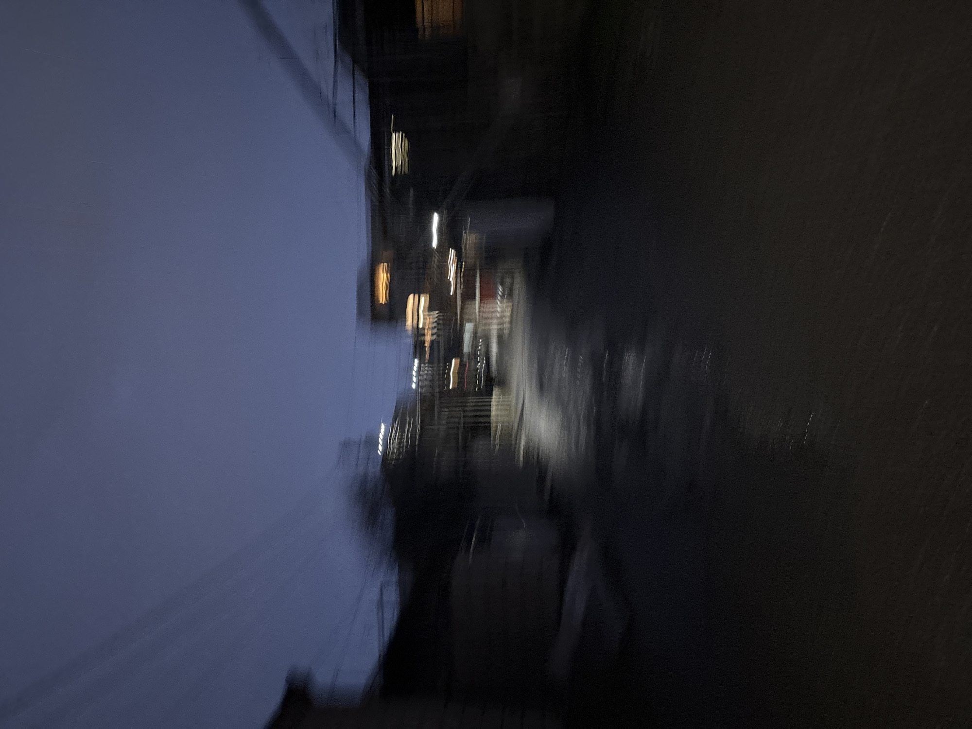The image depicts a blurry, low-light view of a possible urban scene, possibly in Yerevan, Armenia. Visible are indistinct shapes that suggest buildings, structures, and possibly some kind of water feature or dark surface. There are a few illuminated points that are consistent with streetlights. The overall impression is one of motion and low visibility. Due to blurriness, there are not distinguishable people.