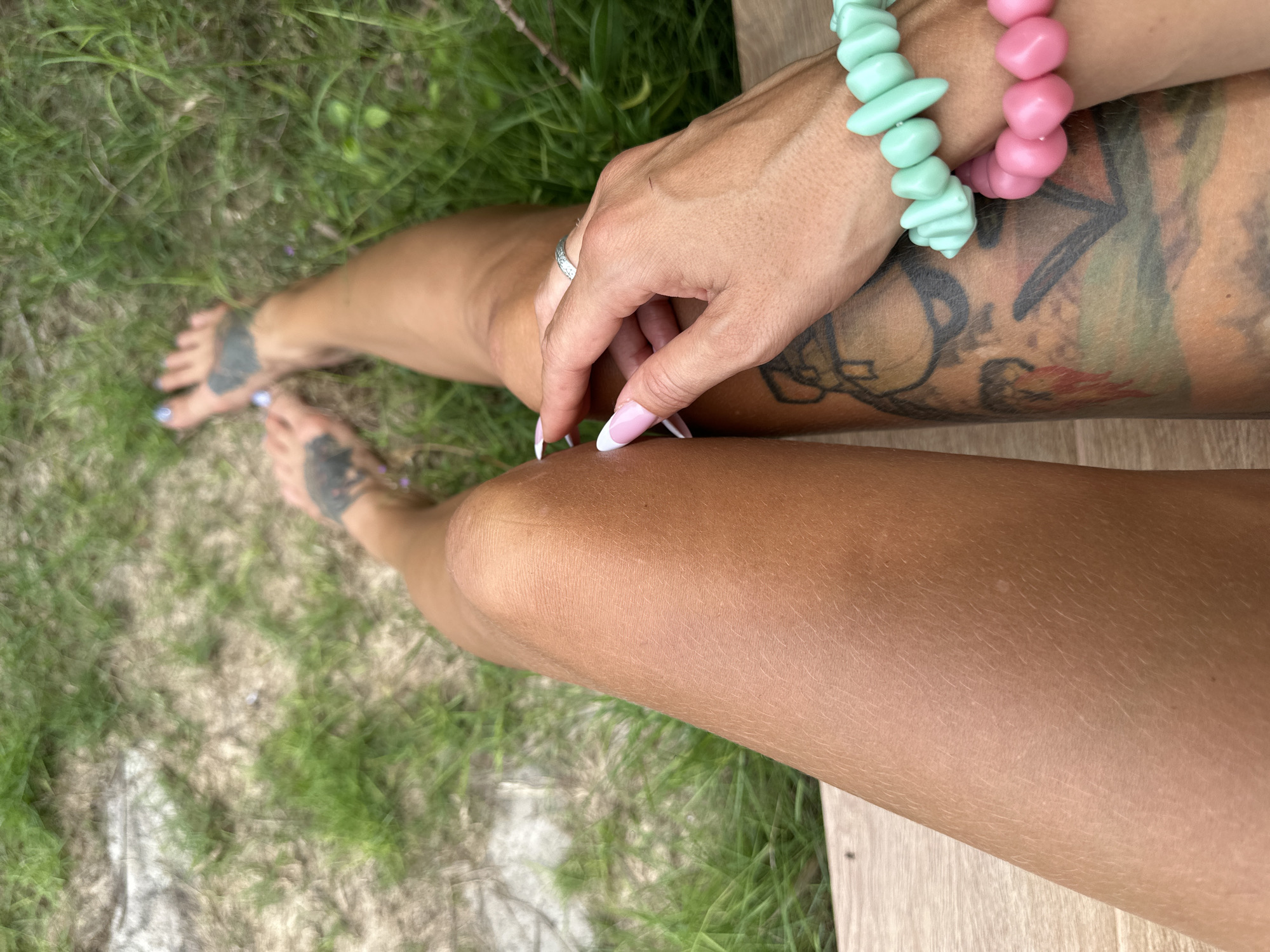 This high-angle, close-up image captures a person's legs, feet, and hands in an outdoor setting. The subject has tanned skin.
One hand, adorned with a silver ring on a finger and French-tipped fingernails (pink base with white tips), rests casually on a bare leg. On the same arm, two distinctive beaded bracelets are worn: one features irregularly shaped mint green or aqua beads, and the other has chunky, round pink beads. The upper part of the leg and arm display colorful tattoos, characterized by abstract or possibly floral designs.
Further back, the person's bare feet are visible, showing light blue or lavender toenail polish and darker tattoos on the top of the feet. The legs are smooth and tanned.
The background consists primarily of green grass, with some patches of dry earth. A light wooden surface, possibly a bench, deck, or boardwalk, is prominent in the foreground and along the right edge, suggesting the person is seated or reclining on it. The bright, even lighting indicates it is daytime, likely sunny. The overall scene exudes a relaxed and comfortable atmosphere in a warm, natural environment, consistent with the specified location of Mũi Né, Vietnam. No text is visible in the image.