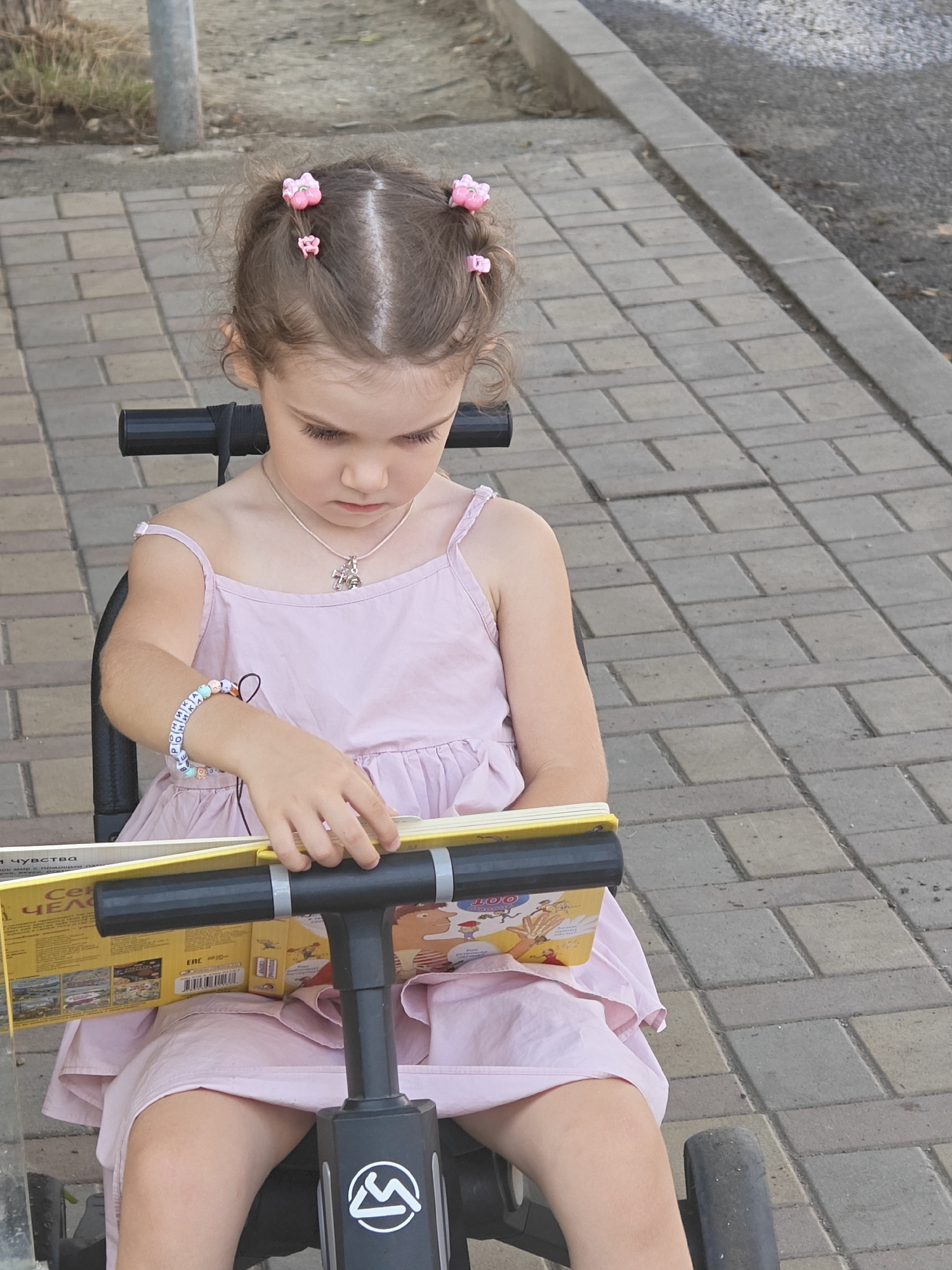 A young girl with brown hair, styled with pink flower clips, sits in a black stroller and looks down at a book. She wears a pale pink dress and a silver necklace with a cross pendant, along with a beaded bracelet on her left wrist. The book she is holding is open and appears to be a children's book with colorful illustrations. The stroller has a black handlebar with silver accents. The girl is positioned on a paved walkway made of rectangular bricks, with a curb and dirt/grass area to the left. The background shows more paved walkway and some dry vegetation.