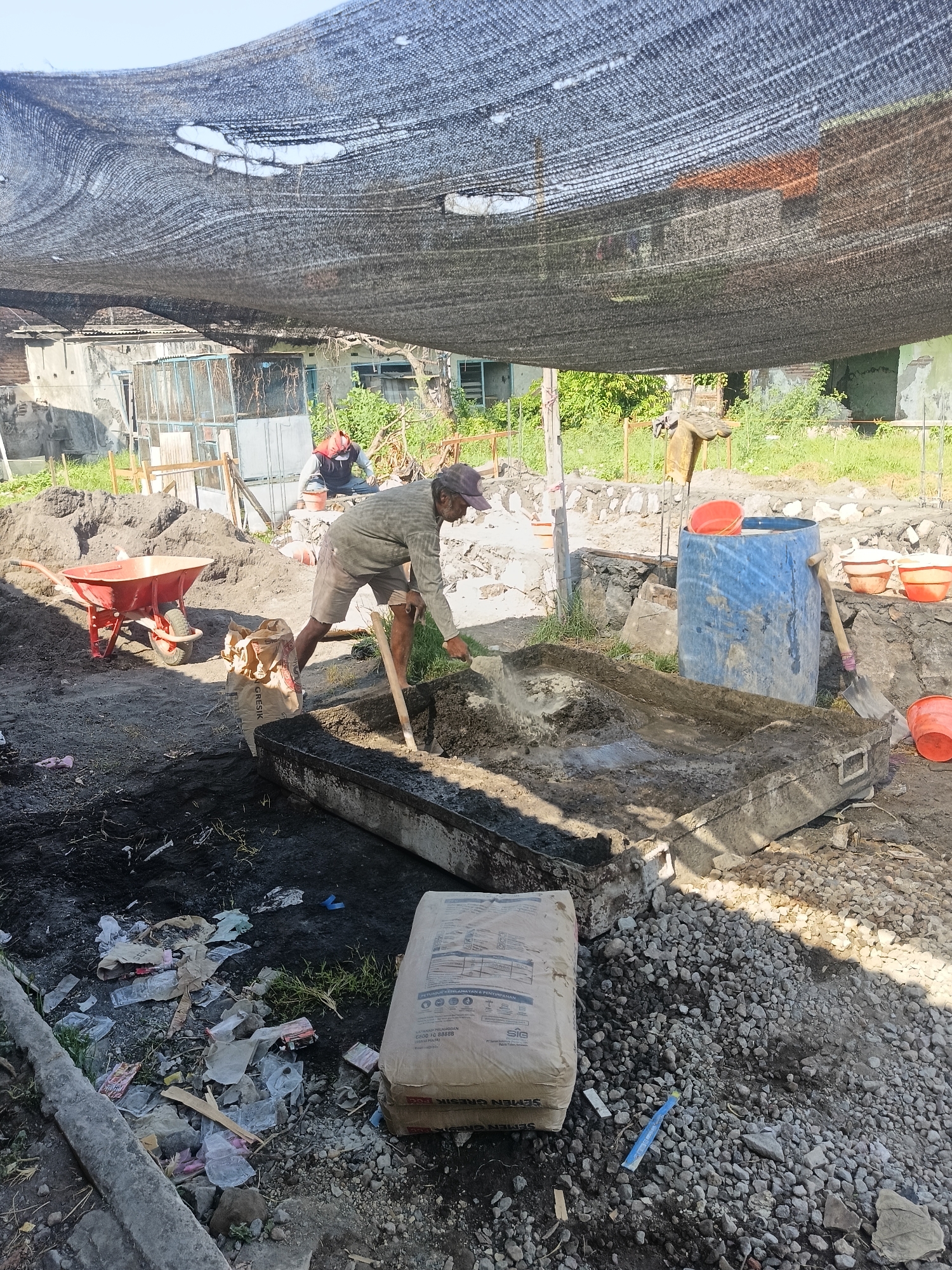 The image depicts a construction site in Gedangan, Indonesia, with a man in the foreground mixing cement by hand using a shovel. He is wearing a light-colored long-sleeved shirt, shorts, and a cap. To his left, a red wheelbarrow is positioned near a pile of sand. A bag of cement lies on the ground in front of him. In the background, another person in a red head covering is also working at the site. The area is partially covered by a black shade net, which is torn in places. Various construction materials, including buckets, a large blue barrel, and rubble, are scattered around the scene. The environment appears to be an outdoor construction area with some vegetation and buildings visible in the distance.