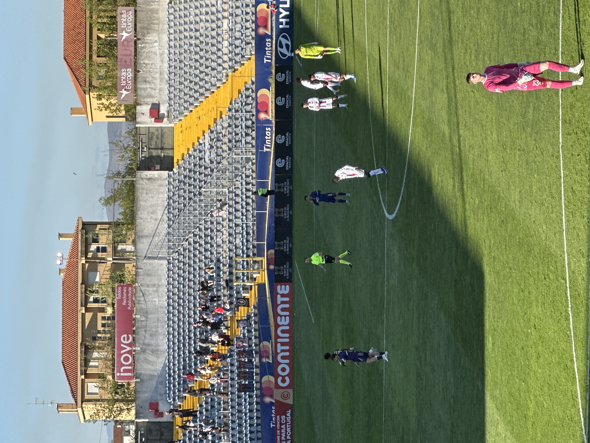 The image captures a soccer game in a stadium in Chaves, Portugal, showcasing several distinct sections.
In the foreground, on the right, a soccer field dominates the scene, populated by soccer players in action. The field shows some shadow.
On the left, stadium seating stretches into the distance, filled with spectators. Buildings are visible, one with a "hove" banner, while another displays "tintas Europa." A large "CONTINENTE" banner and other banner ads like "Tintas" and "HYLI" adorn the stadium infrastructure.