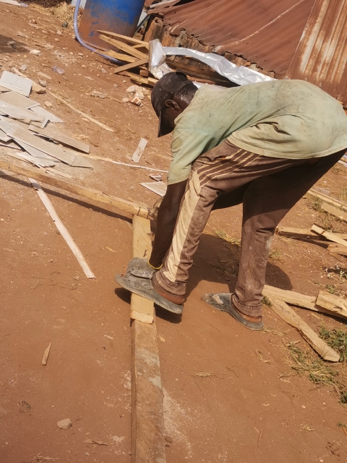 The image captures an outdoor construction or work site in Agbor, Nigeria, on dry, reddish-brown earth. A single adult male is depicted from the back and side, bending forward at the waist. He wears a black baseball cap, a dirty light-green short-sleeved t-shirt, and brown trousers featuring wide vertical tan stripes along the outer seams. Dark sandals are worn over what appear to be light-brown ankle-high socks or foot coverings.
The man's left foot is planted on a long, rectangular wooden beam resting horizontally on the ground. His hands are actively engaged with a smaller piece of wood positioned against the larger beam, suggesting a woodworking task such as shaping or cutting; no specific tool is clearly visible. Sawdust is present on the wooden beam and the surrounding soil near his left foot.
The environment includes scattered wood scraps of various sizes, along with piles of light-colored, thin, flat debris resembling discarded construction materials, particularly on the left side of the frame. In the upper left background, a blue plastic barrel with a white tube or hose extending from it is partially visible. The upper right background features a corrugated metal structure, likely a wall or shed. Sparse, dry vegetation is visible at the edges of the scene.