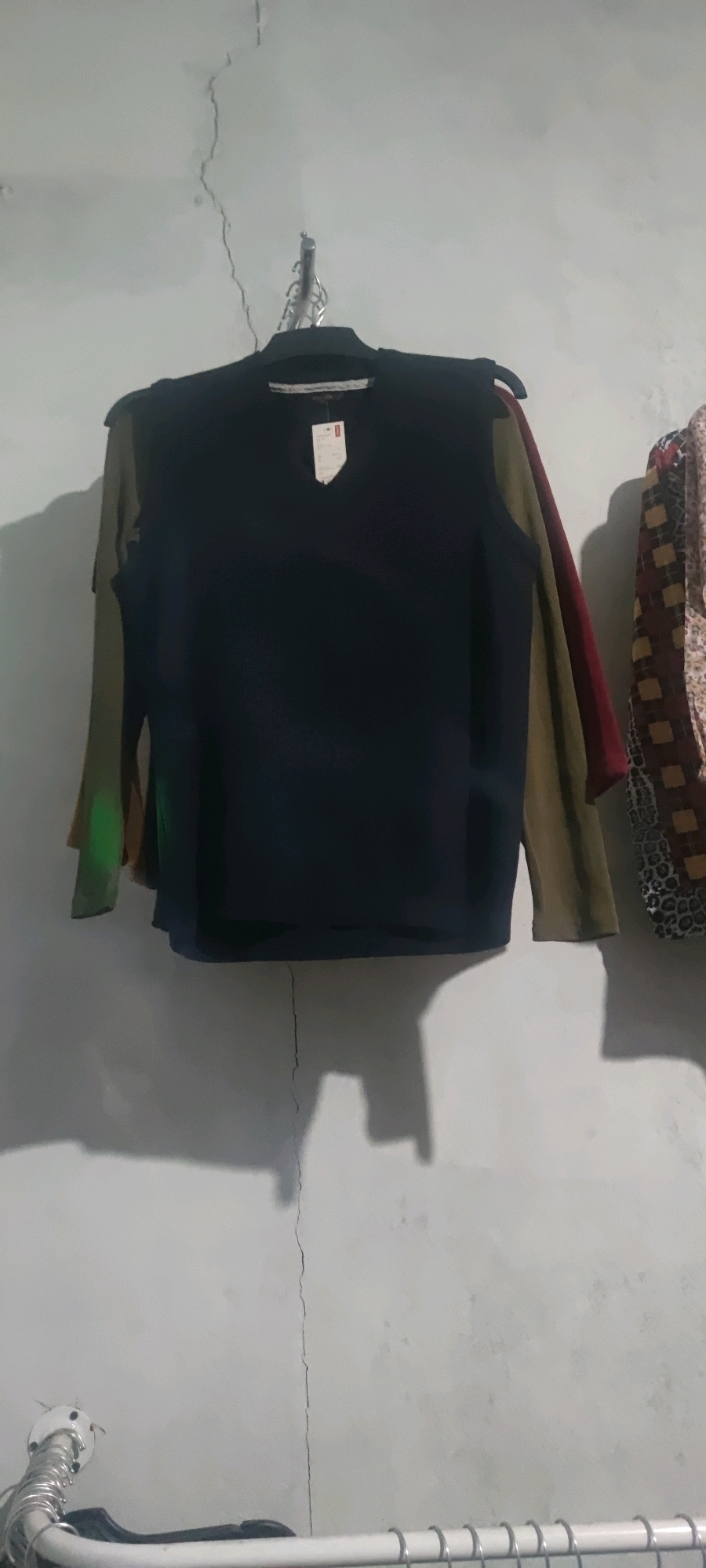 A dark-colored V-neck sweater hangs on a clothes hanger against a light-colored wall. The sweater has long sleeves and appears to be made of a knit material. The wall has a prominent vertical crack running down the center. To the left of the sweater, another sleeve in a dark olive green color is visible, and to the right, a sleeve in a deep red color is visible. Further to the right, a patterned garment with a design of brown and beige squares on a white background is partially visible. The scene is lit from the left, casting shadows on the wall. At the bottom of the image, a metal railing or support structure with numerous circular rings is visible. The context of Gambarsari, Indonesia is not discernible from the visual information provided.