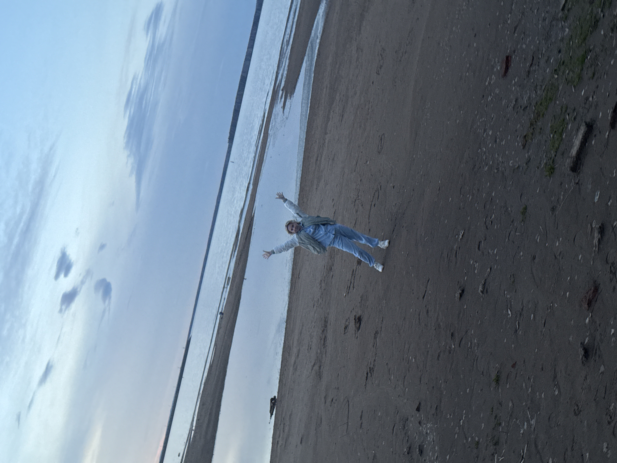 A person is lying on a sandy beach with their arms outstretched, appearing joyful. The beach is situated next to a body of water, possibly a river or lake, under a sky showing some clouds and hues of blue. The location is in or near Chaikovsky, Russia, given the original file name. The foreground of the image reveals a sandy or dirt surface with some scattered vegetation, while the background shows a horizon where the sky meets the water.
