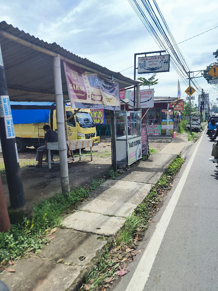 The image shows a street scene in Ciwidey, Indonesia. A man wearing a blue shirt and black pants is sitting on a bench in front of a yellow truck, which is parked on the side of the road. The truck has the words "GUDANG GARAM International" written on its side. There are several food stalls on the right side of the street, some with signs that say "NASI KUTING" and "WARUNG PENANIL." The street is paved and has a white dividing line down the center. There are several motorcycles parked on the side of the road. The sky is clear with a few white clouds. There are power lines running overhead, and the ground is covered in dirt and grass. The street is fairly quiet, with few vehicles or people around.