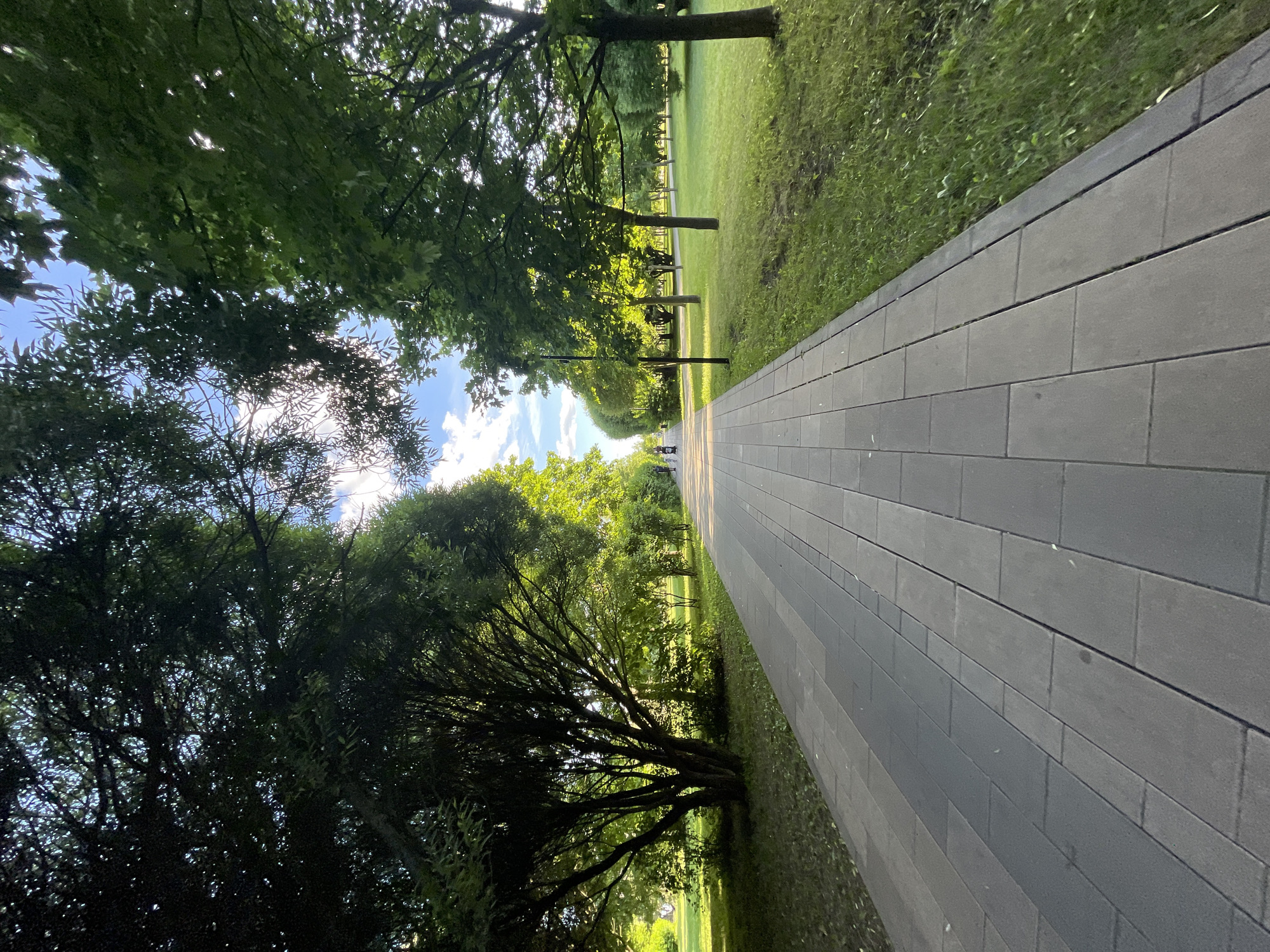 This media file depicts a scenic park path in Moscow, Russia. The scene is dominated by lush green trees casting dappled sunlight on a paved walkway. The path, made of large, grey rectangular pavers, stretches into the distance, bordered by a well-maintained lawn. In the background, more trees line the path, creating a sense of depth and tranquility. The sunlight filtering through the leaves suggests it is daytime, possibly late afternoon given the warm glow. The sky is partly cloudy, with patches of blue visible. A few figures can be seen walking or sitting in the distance, enjoying the park. The overall atmosphere is peaceful and serene, inviting one to explore the park further. There is no visible text in the image.