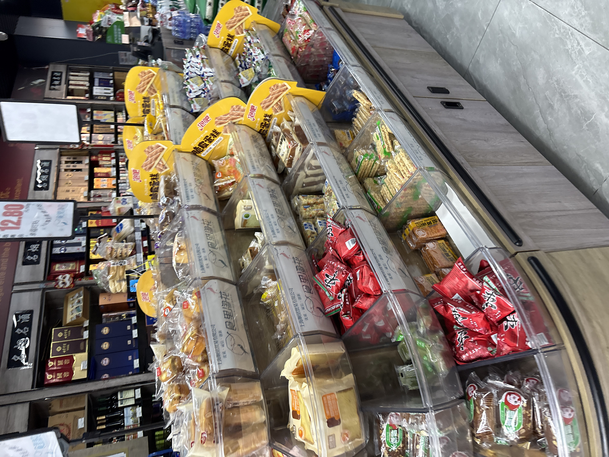 The image depicts a retail display in a store located in Minhang District, China. The display features an assortment of packaged snacks and food items arranged in clear plastic bins. Various types of cookies, pastries, and snack bars are visible, with prominent branding on the packaging. Some items are in red and yellow packaging, suggesting specific product lines. The shelving unit is made of wood-effect material, and the floor is tiled. The overall impression is of a well-stocked convenience store or supermarket aisle.