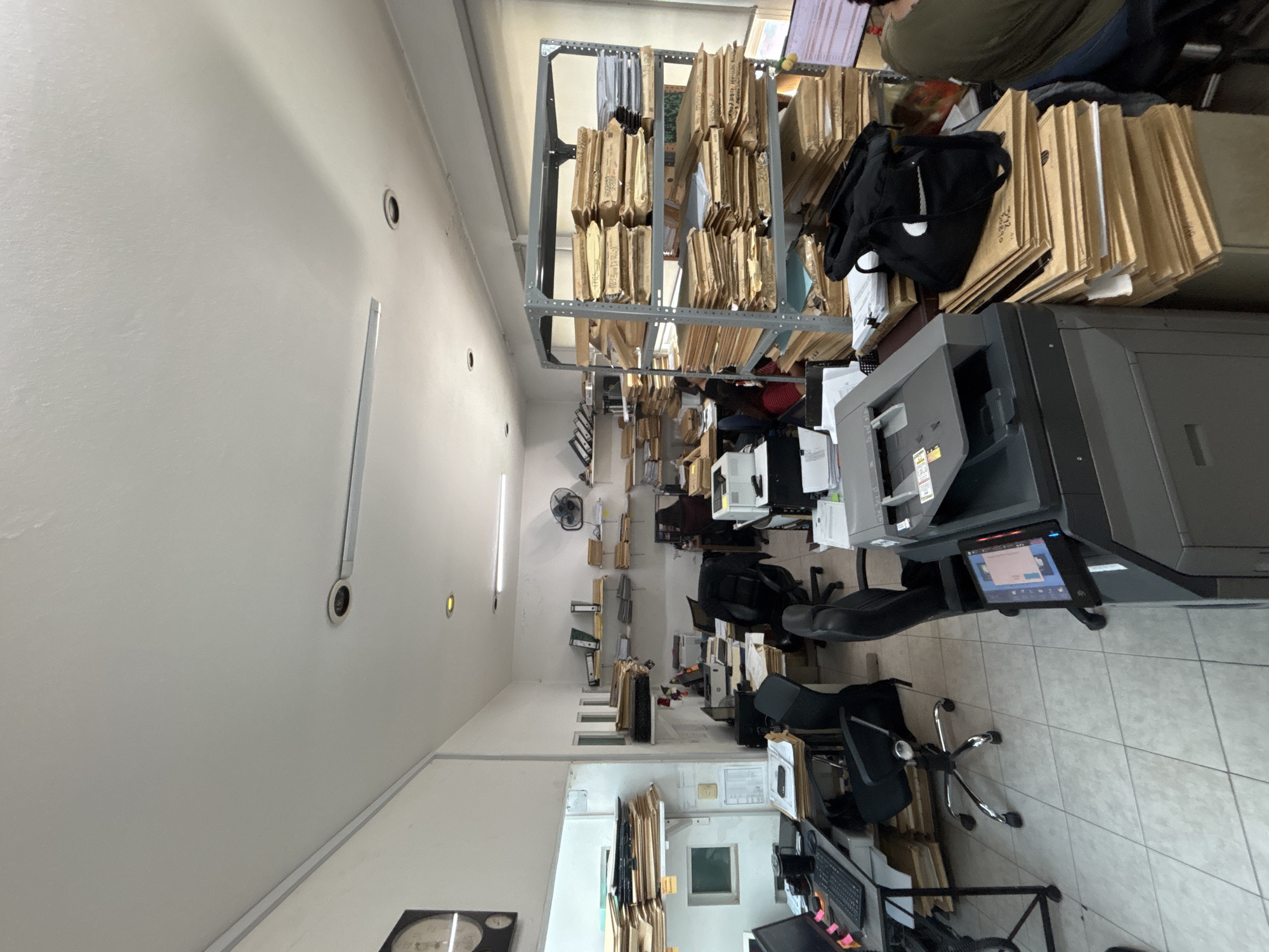 This image captures an indoor office or administrative workspace, characterized by an abundance of physical documents and office equipment. The scene is depicted from a slightly elevated perspective, looking across a room that appears to serve as both an active work area and an archive.
In the foreground and mid-ground, multiple metal shelving units are densely packed with manila folders or brown paper envelopes, indicating extensive record-keeping or document storage. A large, grey multifunction printer or copier is prominently positioned on the right side of the image, with its digital screen displaying a graphical interface. Nearby, a black bag with a distinct white logo (possibly a stylized checkmark or swoosh) rests on a stack of files.
Several office workstations are visible, each featuring computer monitors, keyboards, and ergonomic chairs with wheels. One individual is partially seen from behind in the upper right quadrant, wearing a dark shirt and blue jeans, appearing to be engaged in work at a desk surrounded by files. Further back in the room, additional desks, chairs, and computer equipment suggest multiple active work stations. A ceiling fan, fluorescent tube lights, and recessed lighting fixtures are installed on the white ceiling. The floor is covered with light-colored square tiles.
The overall activity taking place appears to be routine office work, likely involving document management, printing, and data processing. The cluttered yet functional appearance suggests a busy environment focused on administrative or archival tasks. There are no clear time-of-day indicators, as the lighting is artificial. No explicit geographical cues are visible within the image to confirm the provided context of Puebla, Mexico. Text on the numerous file folders is present but too small and indistinct to be legible.