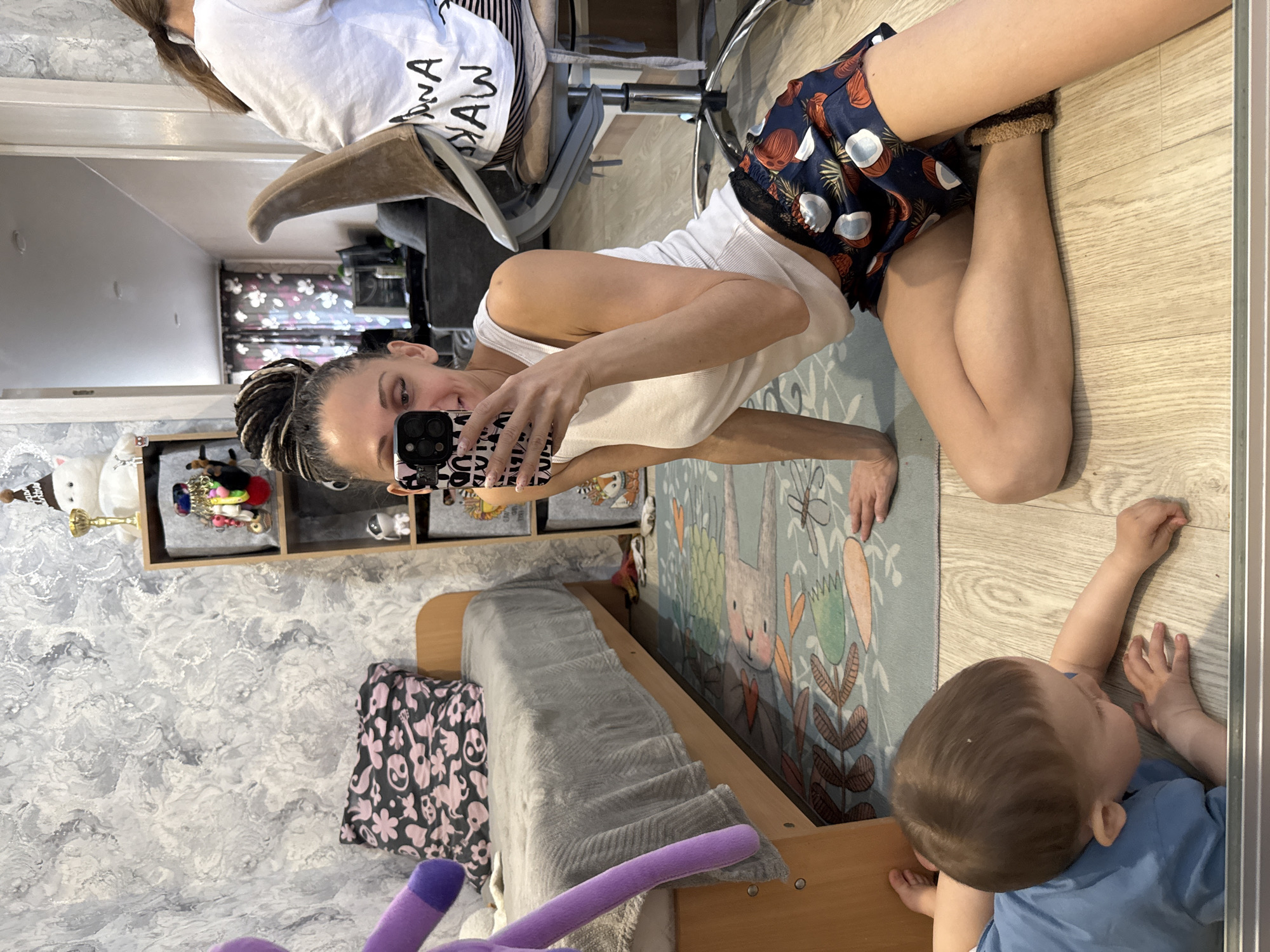 A woman is taking a selfie in front of a mirror, likely in her apartment in Rusanovo, Russia. She is wearing a white tank top and patterned shorts. A child in a blue shirt is lying on the floor near the bottom of the mirror's reflection. In the background, there's a bed with a grey blanket, a patterned pillow, a stuffed purple toy, a rug with a bunny design, shelving with toys and decorations, curtains, and an office chair.