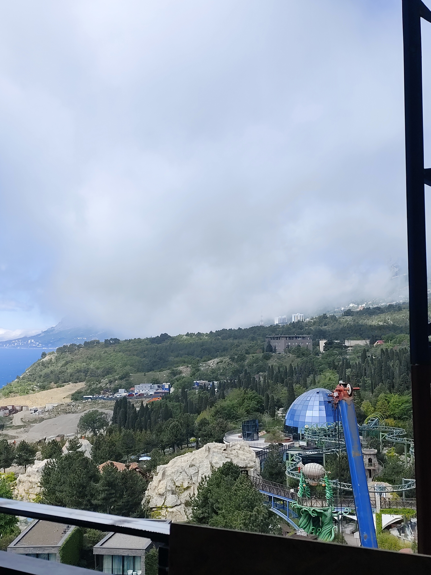 The image shows a landscape with a view of mountains, trees, buildings, and a body of water. In the foreground, there's what appears to be a portion of an amusement park with a roller coaster, a blue dome structure, and various other park features. A crane is positioned near the amusement park structures. In the background, there's a coastline with a mountain range shrouded in clouds. Buildings are visible on the hillside. The scene is observed from an elevated perspective, possibly through a window or from a high vantage point. The sky is overcast. The location appears to be a hilly or mountainous region near the coast.