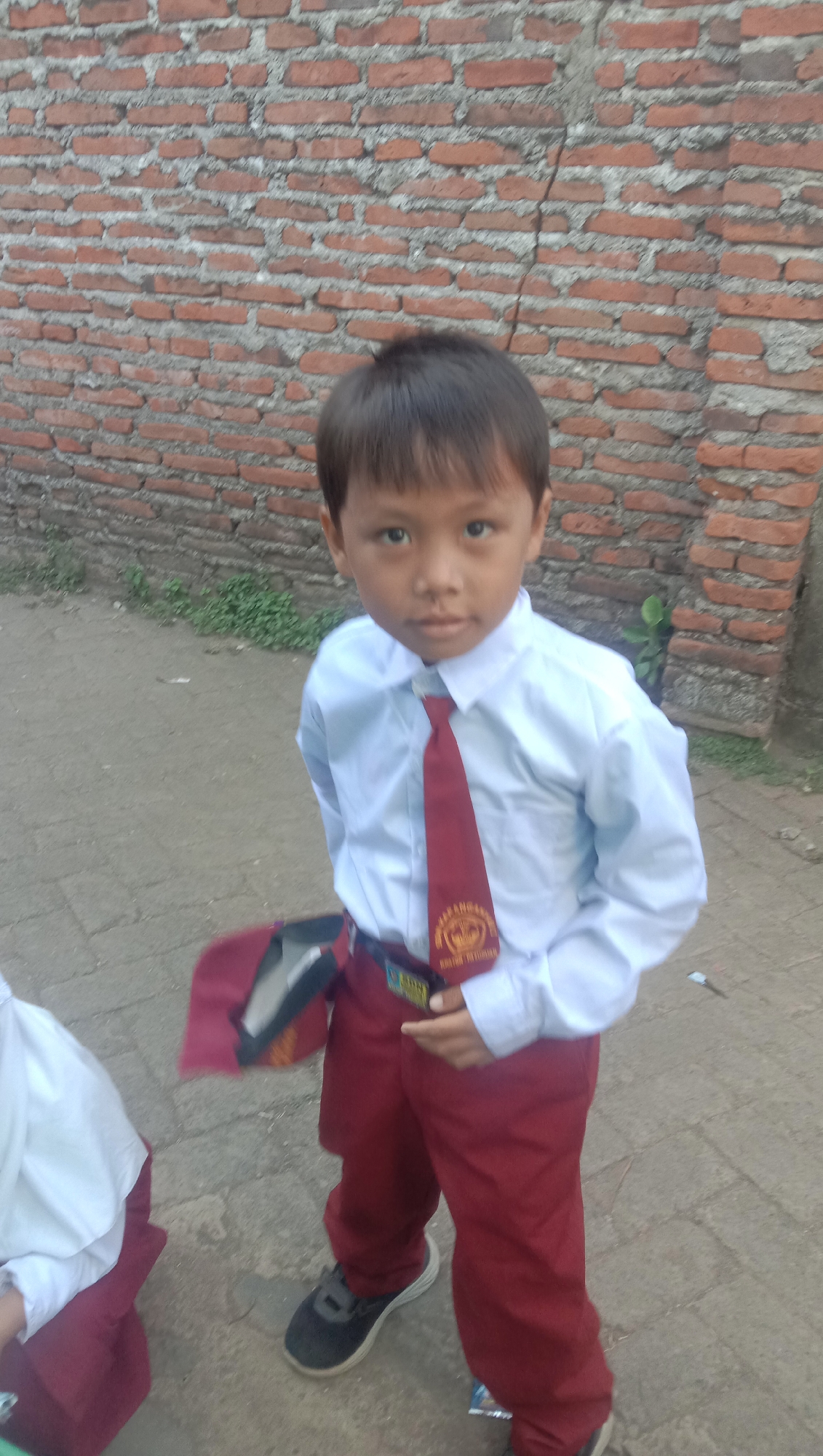 In Karang Anyar, Indonesia, a young boy stands in front of a brick wall. He is wearing a white collared shirt, a red tie with a circular emblem, and maroon pants. His dark hair is cut short. To his left, another child is partially visible, also wearing a white shirt and maroon pants. The ground is paved with what appears to be concrete or asphalt. A maroon bag is draped over the boy's left arm.
