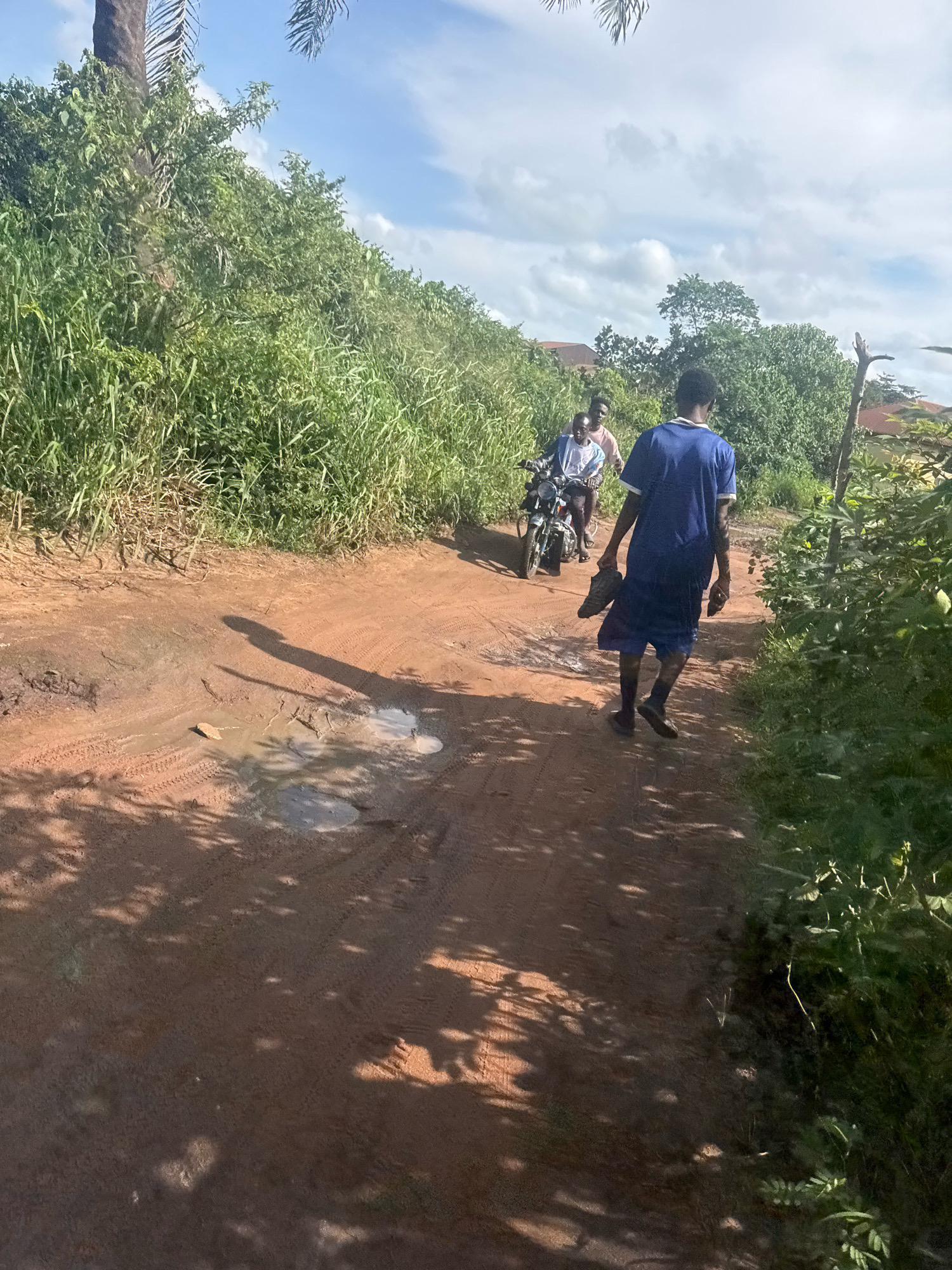 In Ihiagwa, Nigeria, a dirt road passes through lush green vegetation under a partly cloudy sky. A young man, dressed in a blue shirt and shorts, walks barefoot on the sandy road, carrying a pair of dark shoes in his right hand and another shoe in his left hand. Behind him, a motorcycle with two individuals on it follows. The motorcycle rider wears a light-colored shirt, and the passenger behind him wears a pink shirt. The road shows tire tracks and a few puddles of water, indicating recent rainfall. Sunlight filters through the foliage, casting shadows on the ground. Buildings with red roofs are visible in the background, peeking through the dense greenery.