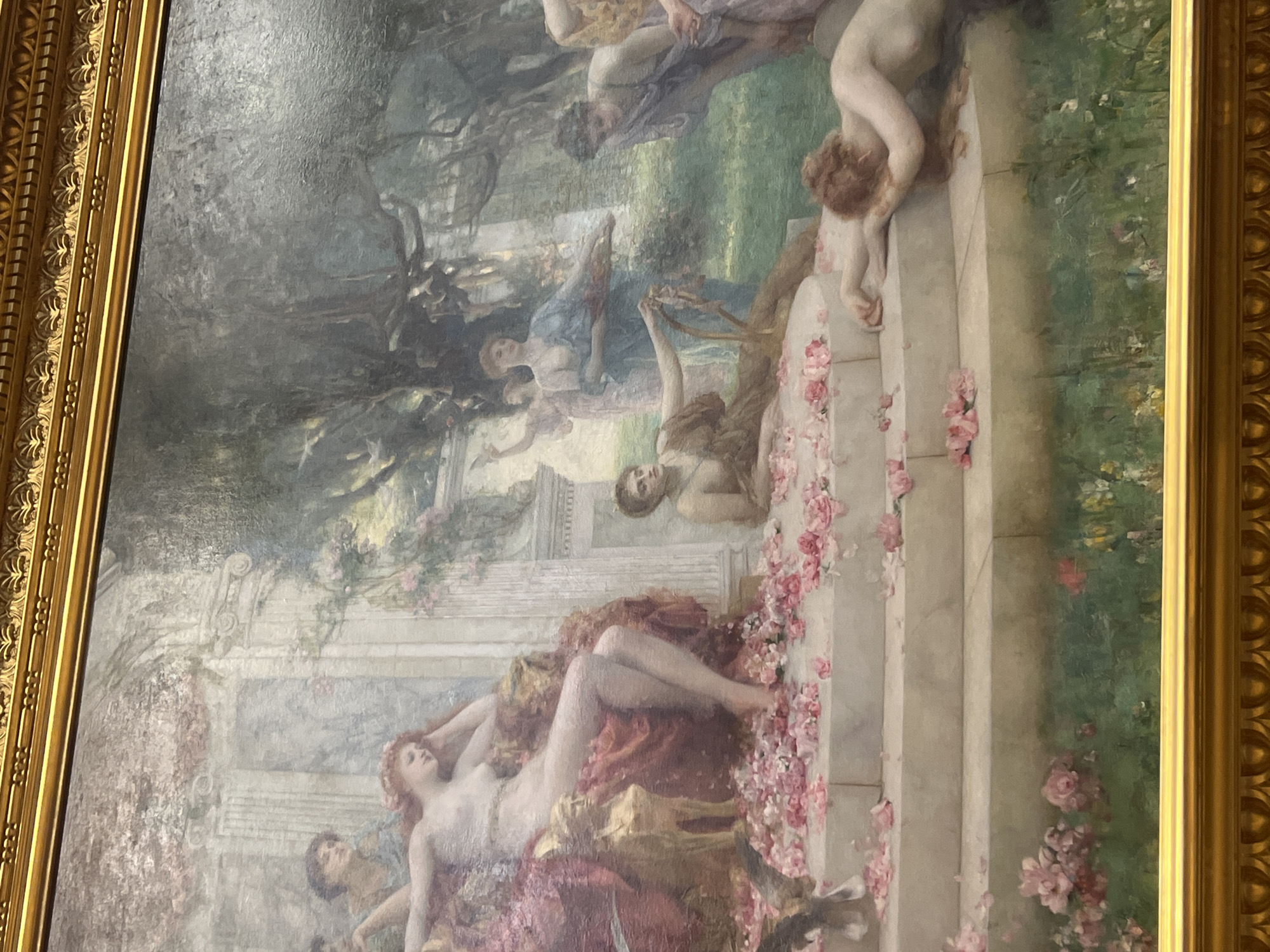 An ornate, gold-framed painting, oriented vertically in the image but depicting a horizontal scene, presents a classical or mythological garden tableau. Numerous figures, predominantly nude or semi-nude women and several cherubic beings, are scattered across a lush landscape featuring trees, foliage, and classical architecture.
A prominent feature is a grand marble staircase or balustrade, running diagonally from the lower right to the mid-left of the painting (when viewed correctly), lavishly adorned with scattered pink rose petals. Several figures interact with or recline upon these steps. In the foreground, a reclining, fair-skinned woman with red-gold hair, almost entirely nude, rests on rich red and gold drapery, gazing upwards. Another cherubic figure with reddish hair is positioned on the steps to the upper right. Other female figures are visible in various poses, some seemingly conversing, others gazing into the distance, with one appearing to hold a stringed instrument like a lyre.
The setting suggests an idealized, possibly ancient, garden with hints of water in the background. The lighting is soft and diffused, indicating daytime, perhaps late afternoon, contributing to an overall serene and idyllic atmosphere. The figures express a sense of calm, beauty, and sensual leisure. No discernible text is visible within the artwork. The heavy, gilded frame features intricate carvings. The painting is located in London, United Kingdom.