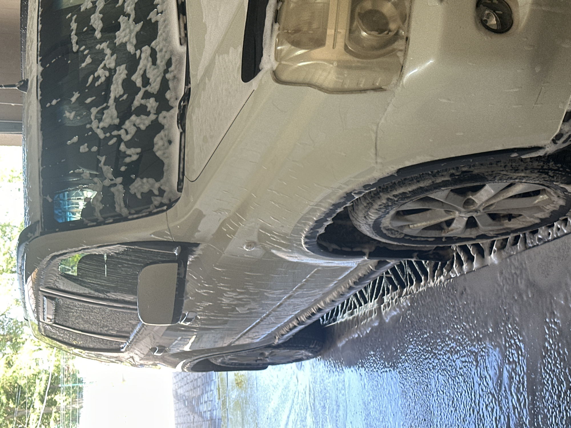 The image shows a white SUV being washed at a self-service car wash. The vehicle is covered in soap suds, and the ground is wet and foamy. The car wash appears to be outdoors, with trees and foliage visible in the background. The scene is brightly lit, suggesting it is daytime. The specific location in Shatava, Ukraine, is not discernible from the image itself, as no identifying landmarks or signs are present. The image is a close-up of the front passenger side of the vehicle, showing the headlight, fender, wheel, and part of the door. The car is positioned over a grate, which is used to drain water and cleaning solution.