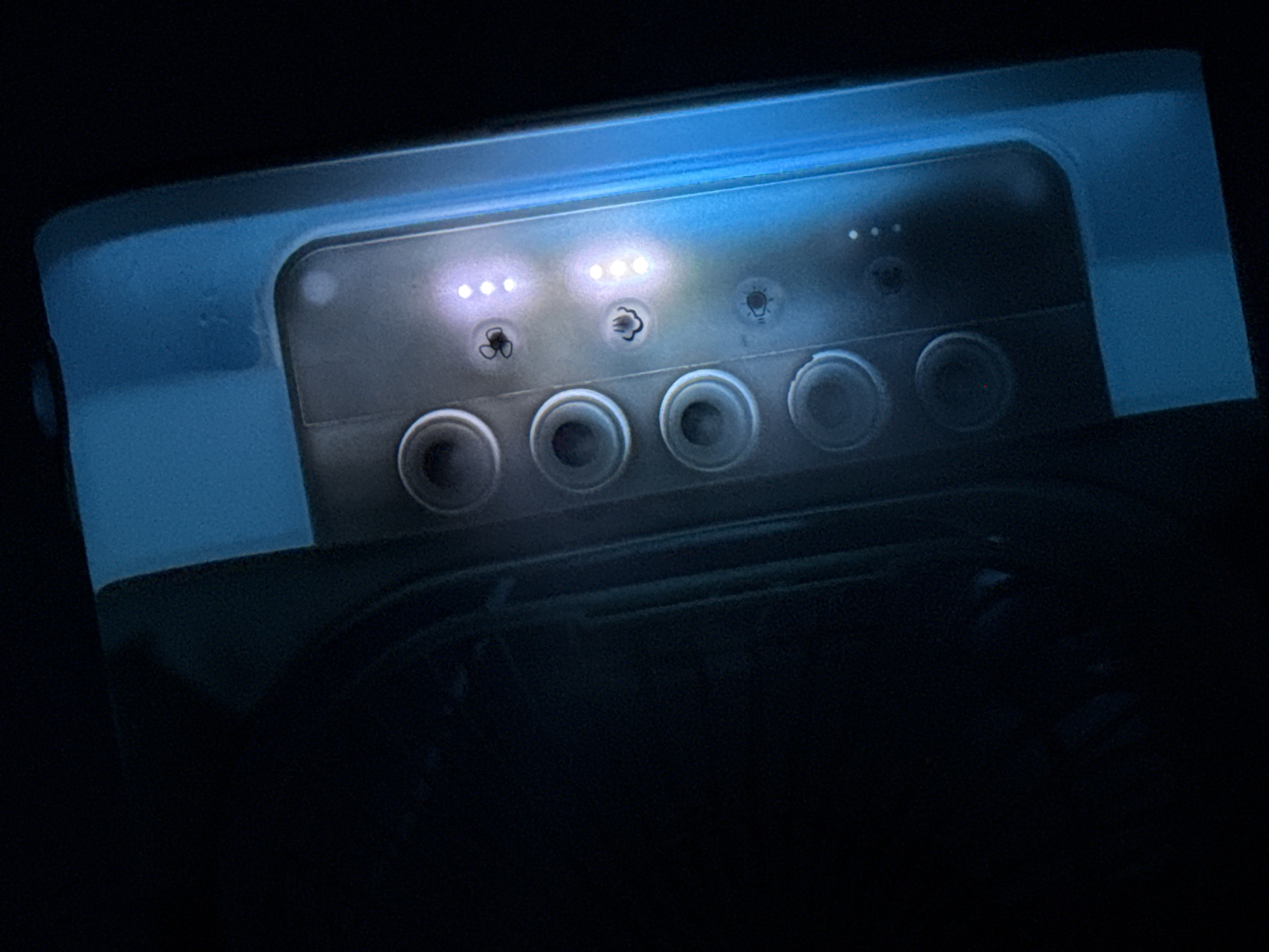 The media file displays a close-up of an illuminated control panel or interface in a dark environment. The scene is dominated by the blue-tinted device, which features a row of indicator lights and symbols above a row of circular controls.
In the upper section of the panel, there are several small, circular indicator lights. On the left, a group of three lights shows two illuminated. Next to it, another group of three lights is fully illuminated. Below these lights are two distinct symbols: the first resembles a fan, and the second appears to be a puff of smoke or steam. Further to the right, a symbol resembling a light bulb is visible, with a single, unlit indicator light above it.
Below the illuminated indicators and symbols, there are five prominent circular objects arranged in a row. These appear to be buttons, dials, or input controls for the device. The entire control panel is set against a dark, possibly black, background, emphasizing the bright, cool-toned illumination of the device itself. The primary colors in the image are various shades of blue and white from the device's lighting, contrasting with the deep black of the surroundings. The time of day is not discernible, but the deep shadows suggest a low-light condition. No visible text is present on the panel.