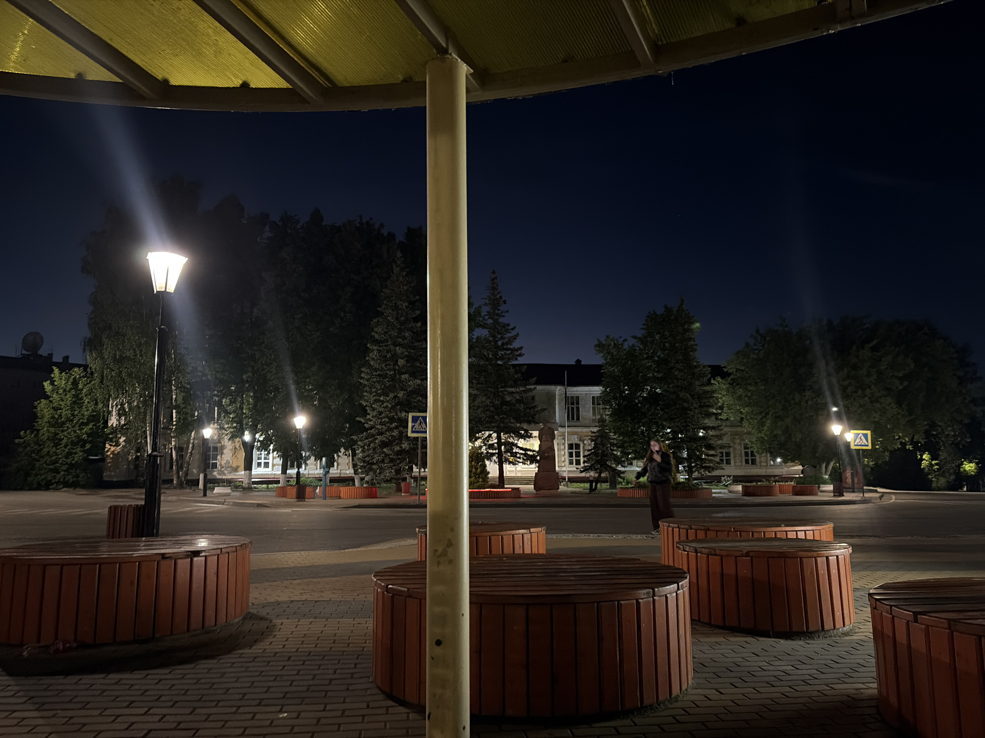 The image depicts an outdoor scene at night, under a yellow canopy supported by a central pole. In the foreground, several circular wooden benches with a reddish-brown hue are visible, arranged on a brick-paved ground. A street lamp with a bright light illuminates the area.
In the background, a street with asphalt pavement is visible, lined with trees on either side. Across the street, there are buildings with windows, suggesting an urban setting. A pedestrian crossing sign with a yellow diamond shape and a black pedestrian silhouette is mounted on a pole. Further in the background, a person is standing on the sidewalk near a building. The sky is dark blue, indicating nighttime.
The location is identified as the city of Semyonov, Russia, specifically on Sovetskaya Street, in front of the Semyonov City Palace of Culture. The circular wooden benches and the distinctive yellow canopy are characteristic features of this public space, often used for outdoor events or as a resting area. The presence of street lighting and the building in the background confirm the urban environment.