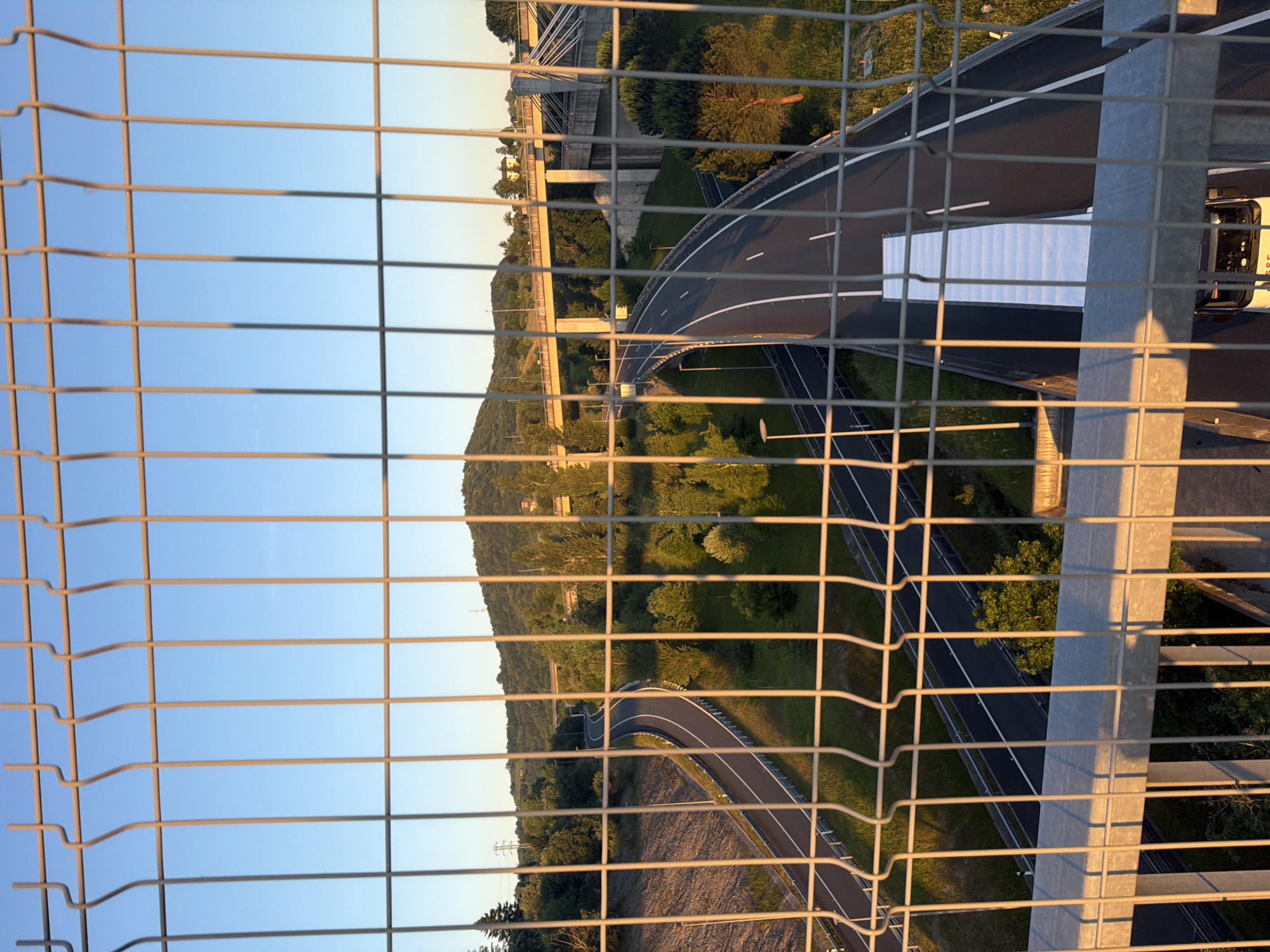 The image shows a view through a metal wire fence in Errenteria, Spain. Visible beyond the fence is a multi-lane highway winding through a green, hilly landscape. A white truck is on the highway. Above the highway are elevated bridge structures. The sky is light blue, indicating daytime, with a hint of sunset colors near the horizon.