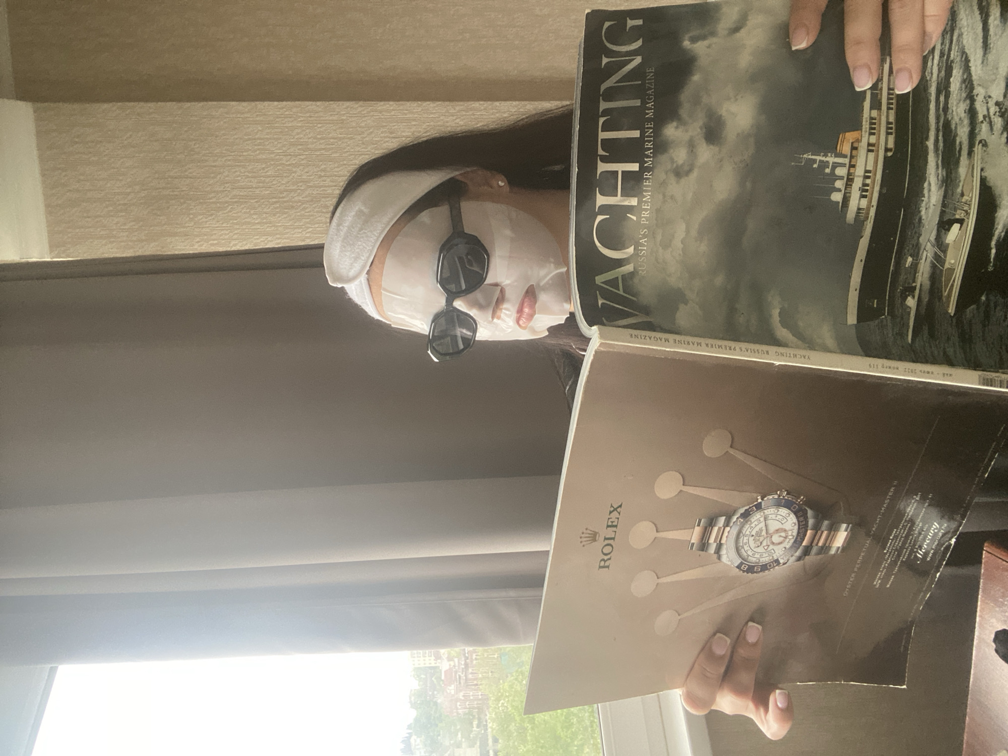 A person wearing a white face mask, sunglasses, and a white headband is holding two magazines. The magazine on the top is "Yachting Russia's Premier Marine Magazine", with the bottom magazine showcasing a Rolex watch. The scene appears to be indoors with a window slightly visible in the background. Based on the magazine's title, the environment is in Russia. The text on the Yachting magazine suggests it could be the 2012 issue. The city of Sochi is not explicitly mentioned or identifiable in this image.