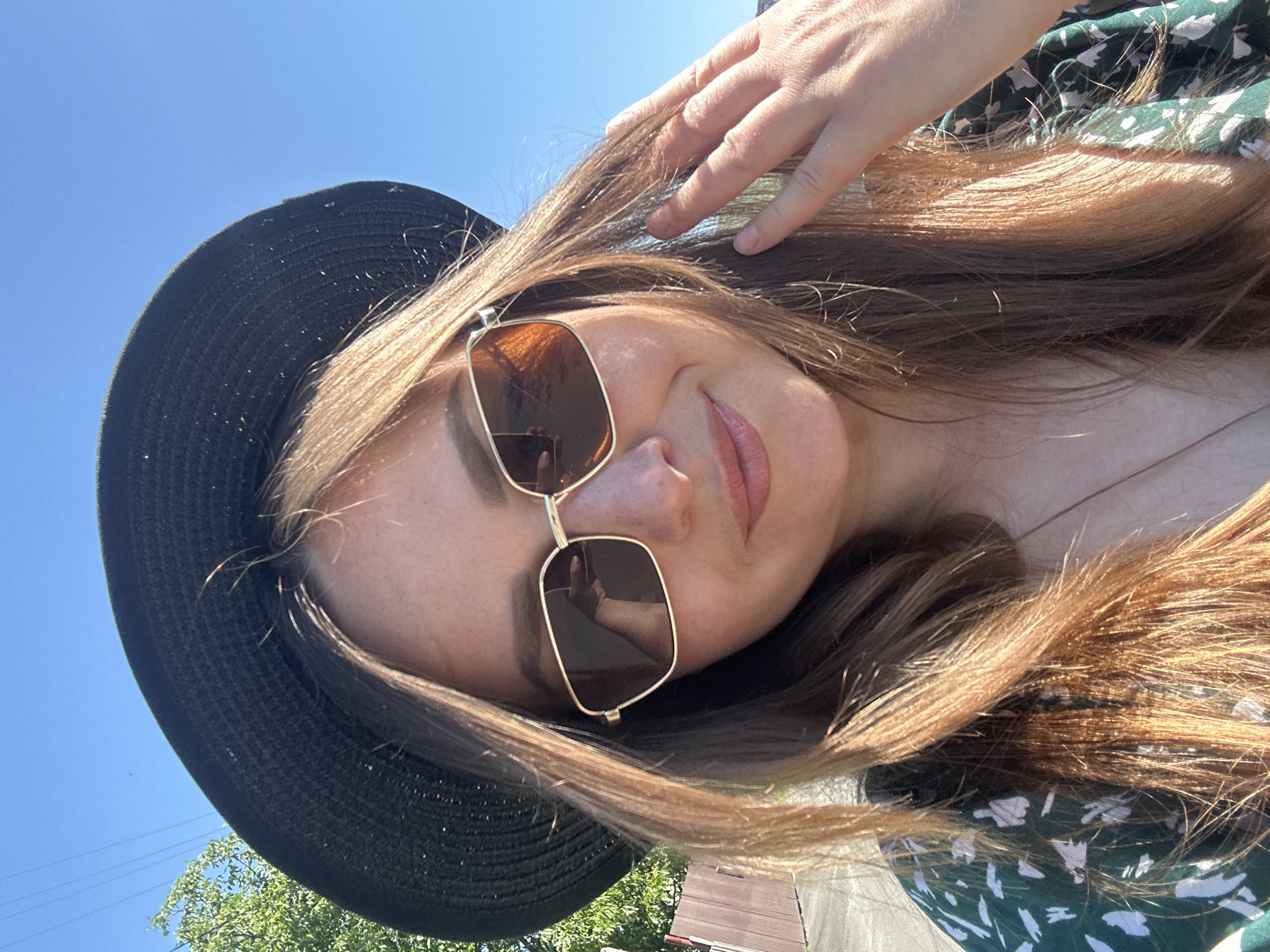 In a selfie taken outdoors, a woman wearing a black wide-brimmed hat and gold-rimmed sunglasses smiles at the camera. Her light brown hair is styled loosely around her face and shoulders, with strands illuminated by sunlight. She is wearing a green dress with a floral pattern. Her right hand is raised, with fingers gently touching her hair. In the background, a clear blue sky is visible, along with the green foliage of a tree and a wooden fence. The lighting suggests it is a sunny day. There are no recognizable landmarks to indicate the specific location within Bila Tserkva, Ukraine.