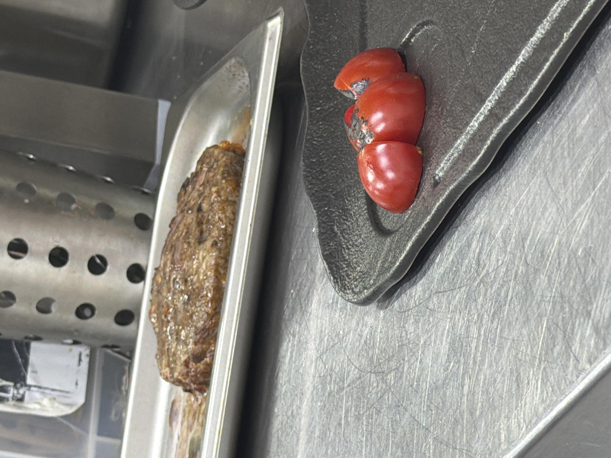 A close-up, eye-level shot shows a piece of cooked steak in a metal tray in the background and three cherry tomato halves on a black plate in the foreground. The steak is well-done with a seared crust. The tomatoes are cut in half and appear slightly charred on top. The scene takes place on a stainless steel countertop, likely in a kitchen.