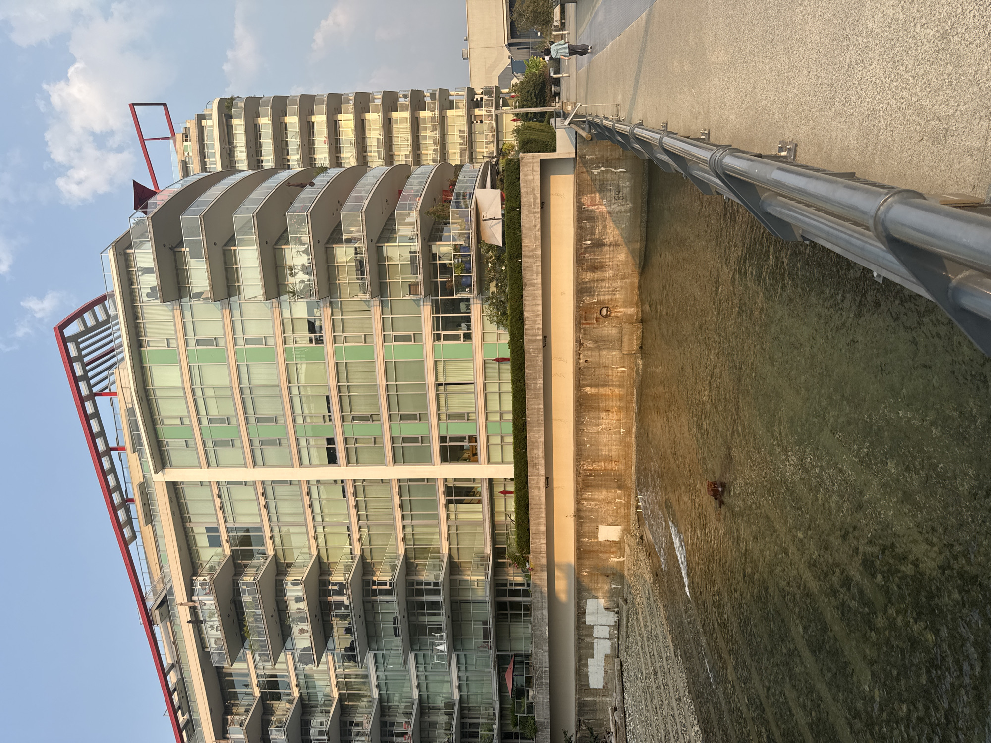 This image depicts a waterfront view in North Vancouver, Canada, likely near the Burrard Inlet. In the foreground, a paved walkway runs along a body of water, bordered by a metal railing. The water itself is shallow and appears to be a riverbed or inlet, with visible stones and some ripples. A small, dark object, possibly a piece of debris or a small animal, is present in the water.
Dominating the background are modern, multi-story apartment buildings with extensive glass balconies and distinct red architectural accents on the rooftops. These buildings exhibit a contemporary design with layered balconies and numerous windows, suggesting a residential or mixed-use development. Greenery is visible on some of the lower balconies and along the waterfront edge.
To the right of the walkway, a person is walking away from the camera, dressed in casual clothing. The sky above is a pale blue with some scattered clouds, and the late afternoon or early evening sun casts a warm, golden light on the scene, highlighting the architecture and the water's surface. The overall impression is one of urban development integrated with a natural waterfront setting.