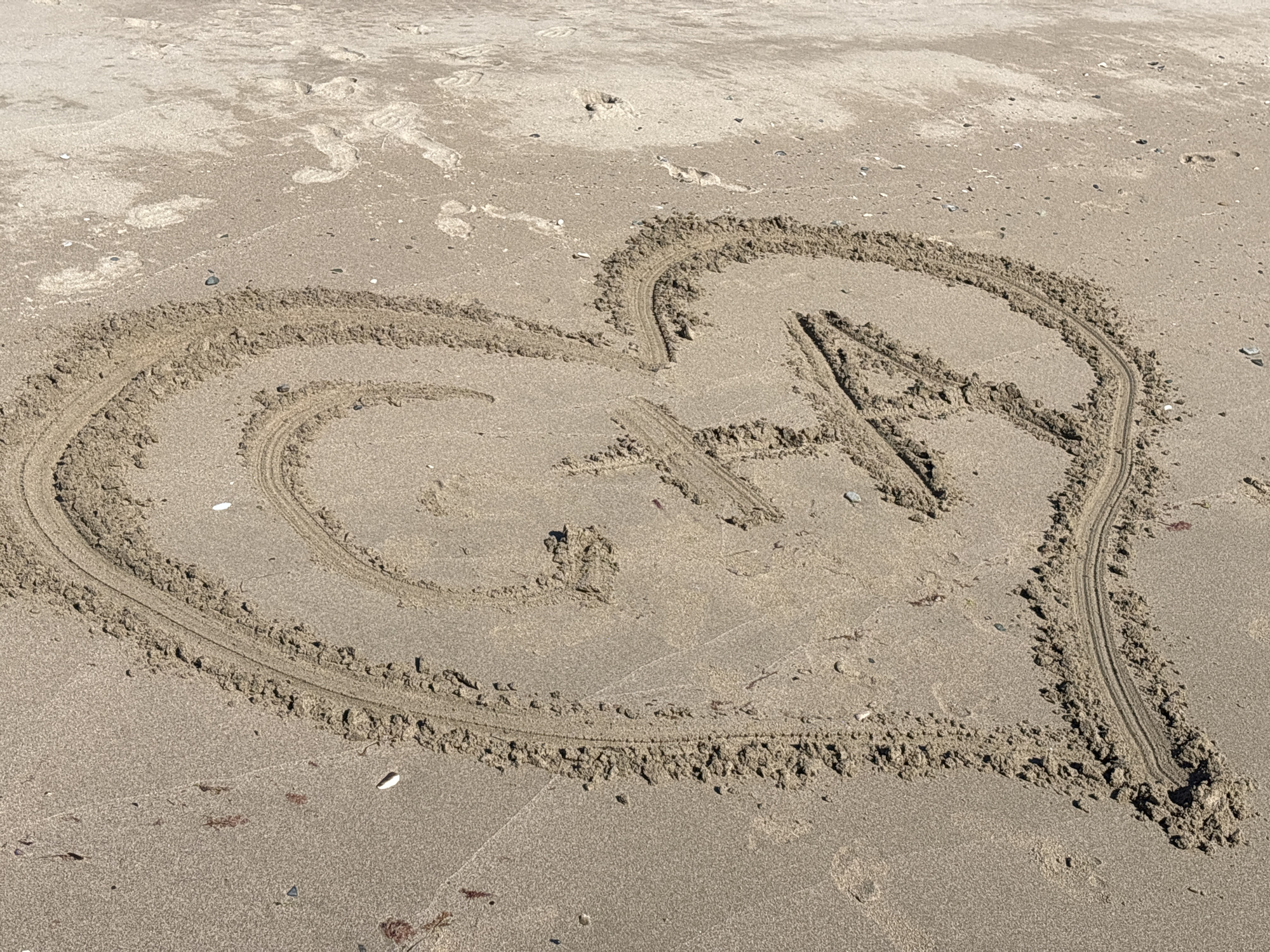 In Rezovo, Bulgaria, a heart shape is drawn in the sand on a beach. Inside the heart, the letters "G" and "A" are also drawn. The sand is light brown and textured, with some small shells and pebbles scattered on the surface. The sunlight creates shadows, indicating a bright, sunny day. The area surrounding the heart appears to be undisturbed sand, with some faint footprints in the distance.