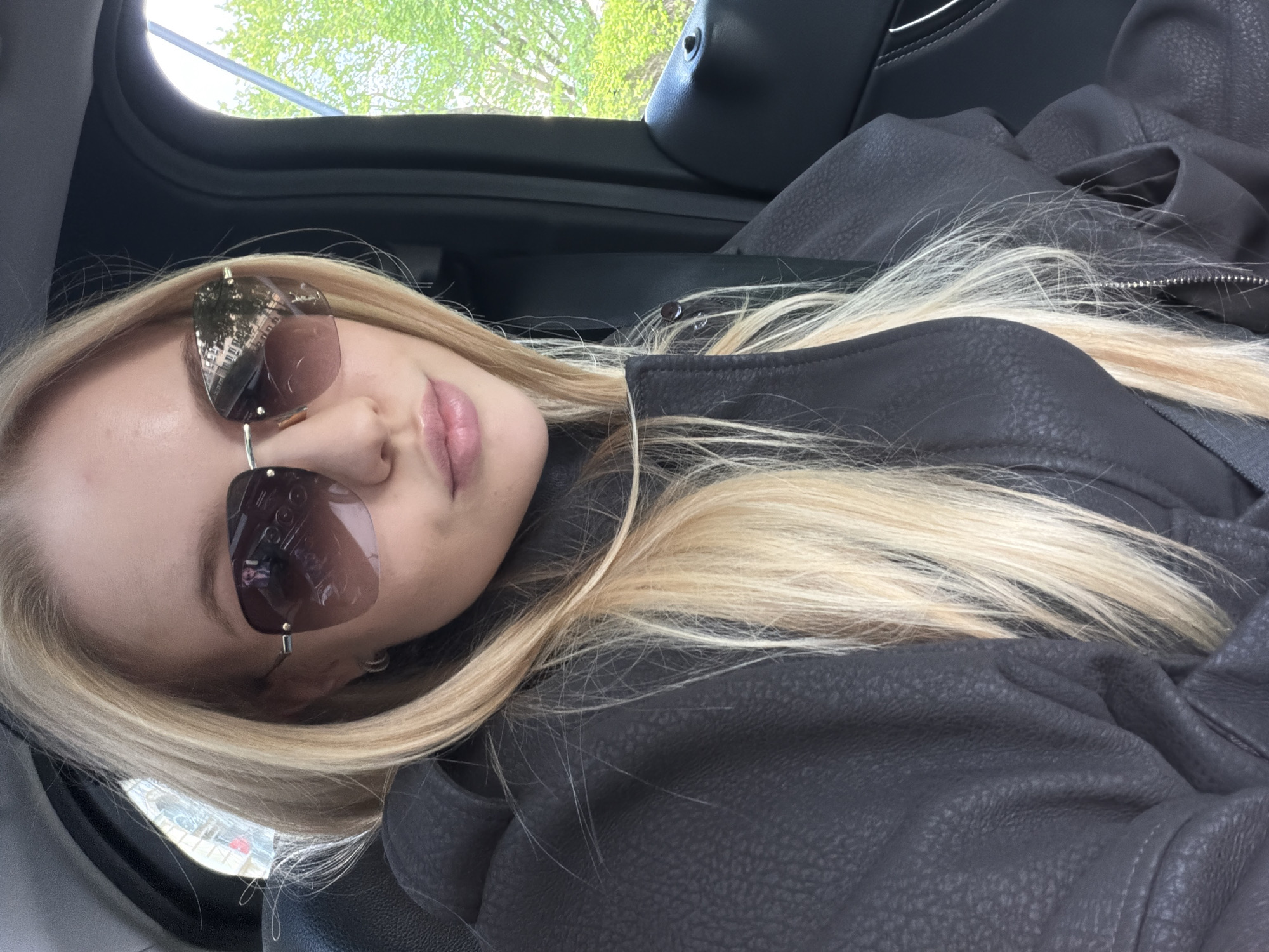 A fair-skinned woman with long blonde hair is shown in the image. She is wearing sunglasses and a dark-colored jacket. The woman is inside a car, as evidenced by the car interior and the window visible in the background. The reflection in her sunglasses indicates a cityscape. The location is in the city of Unknown, Unknown.
