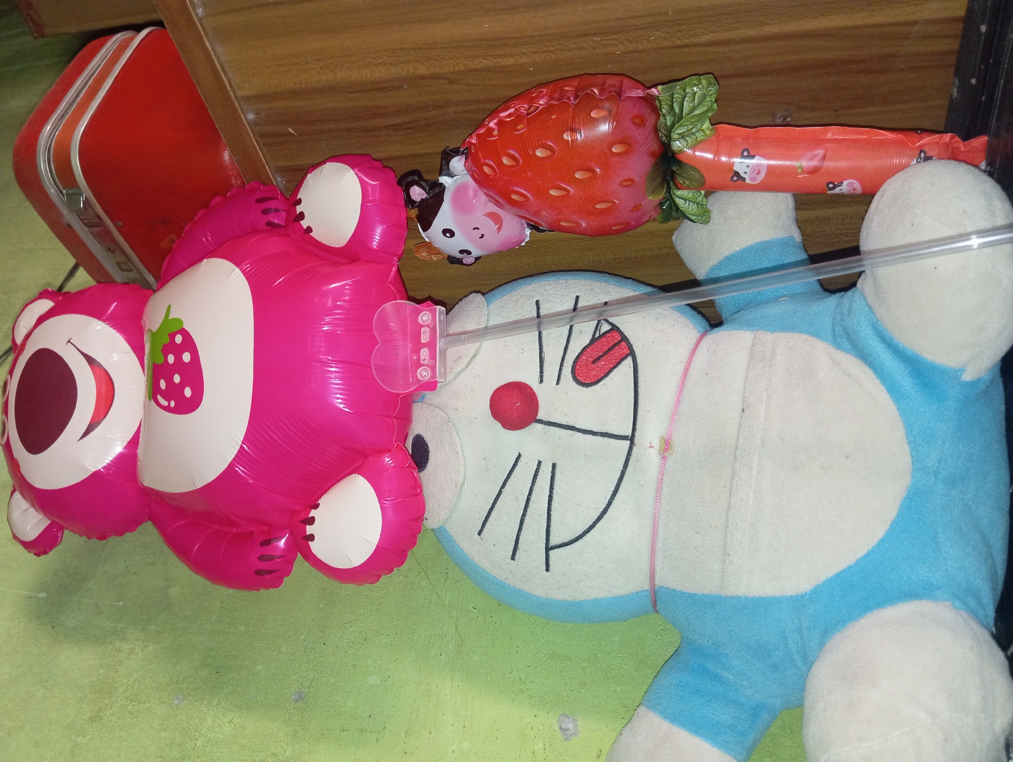 The image shows a collection of stuffed toys and balloons indoors, with a green textured wall in the background and a wooden surface on the right. Dominating the frame is a large, plush blue and white stuffed toy resembling the character Doraemon. To the left of Doraemon, a large pink inflatable balloon shaped like Lotso, the strawberry bear from Toy Story, is positioned above Doraemon. Attached to Lotso's balloon is a clear plastic tube with a pink heart-shaped connector. To the right of Doraemon, a smaller balloon is shaped like a strawberry with a cow's head peeking out from behind it. This strawberry balloon is attached to an orange, elongated object decorated with cow faces and small strawberry graphics, also connected to a clear plastic tube. A red rectangular object, possibly a suitcase or box, rests on the wooden surface behind Lotso. A thin pink string is tied around Doraemon's neck. The lighting in the scene is dim, casting some shadows. The overall impression is a whimsical arrangement of toys and decorative items. The image does not provide any information about the city of Rejosari, Indonesia.