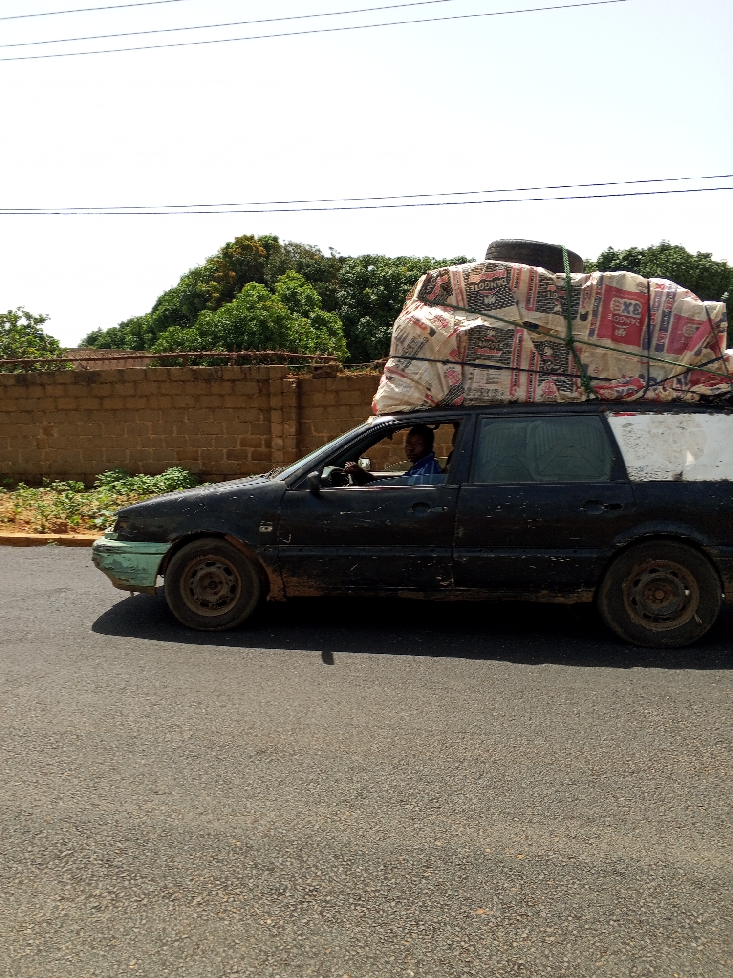 A black station wagon is driving on a paved road. The car is carrying a large load of bagged cement on its roof, secured with ropes and a spare tire. A man is visible driving the car. The car is old and appears to be in disrepair, with dirt and debris on its exterior. In the background, there is a brick wall and lush green trees. The sky is bright and clear. This scene is located in Bukuru, Nigeria.