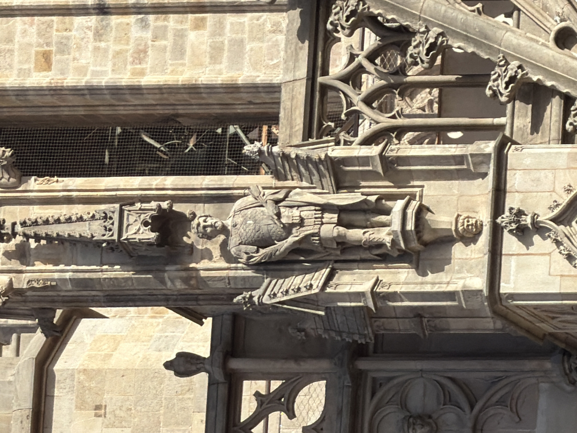 The image shows a close-up of the exterior of a Gothic-style building in Barcelona, Spain. The primary focus is a stone statue of a man in armor, standing on a pedestal. The man has a beard and is wearing a tunic with armor plating over it, a cape draped over his left shoulder, and boots. He is holding a scroll or book in his left hand. The statue is positioned on the corner of a building, with ornate architectural details surrounding it, including spires, carved stone elements, and arched windows with a grid-like covering. The stonework of the building is light-colored, possibly sandstone, and shows signs of age and weathering. The lighting suggests it is daytime, with strong shadows indicating sunlight. The background includes more of the building's stone facade, a large window with a dark grid, and other architectural embellishments.