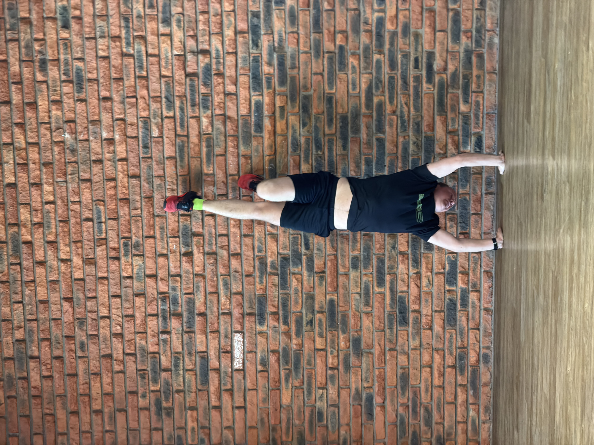 An adult male is captured in a full-length, vertically oriented photograph, although the image itself is rotated 90 degrees counter-clockwise. He appears to be performing a challenging exercise, holding his body horizontally, parallel to the ground, with his back against a textured brick wall. His left hand is pressed against a lighter wooden surface, which would be the floor if the image were correctly oriented, and his feet are pressed against the brick wall.
The man is wearing a short-sleeved black t-shirt with an unidentifiable green logo or text on the left chest area, black athletic shorts, and athletic shoes. One shoe is predominantly red, while the other is black with green accents. He also wears a dark-colored wristband or watch on his left wrist. He has short dark hair and a beard or stubble, with a focused and neutral expression during the exercise.
The background consists of two distinct surfaces meeting at a corner. The prominent surface is a rustic brick wall, composed of various shades of reddish-brown and darker, almost black, bricks, held together by light gray mortar. The bricks vary in size and texture, giving the wall a rugged appearance. The other surface, to the right of the man, is a light-colored wooden paneling or floor, exhibiting a distinct wood grain pattern. The lighting is bright and even, suggesting an indoor setting. There are no overt visual cues in the image itself to confirm the location, but it is contextualized as Chaikovsky, Russia.