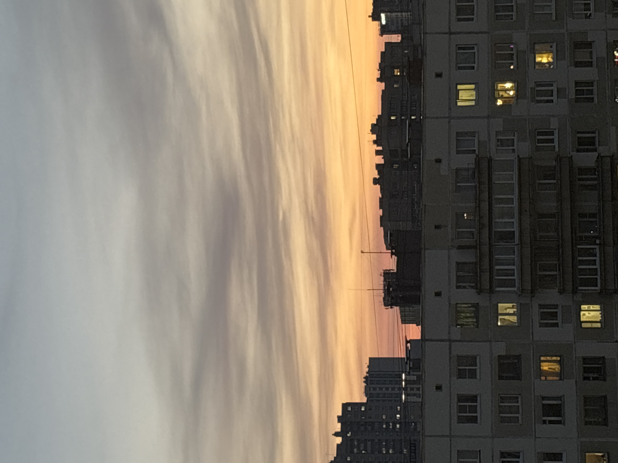 The image presents a view of a cityscape at dawn or dusk. Predominantly, it captures a dense array of apartment buildings, likely in Vsevolozhsk, Russia, characterized by their uniform, box-like structures. Some apartment windows are illuminated, suggesting occupancy and varying activity inside. A gradient sky transitions from gray to orange, providing a backdrop and defining the lighting conditions. Overhead wires stretch across the skyline, contributing to the urban landscape. There is no visible human presence or direct interaction between elements within the scene; instead, the image focuses on the static environment.