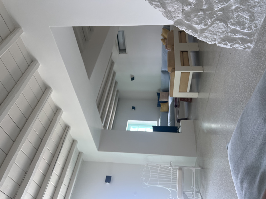 This image displays the interior of a residential living space, likely in Tourlos, Greece. The room features a minimalist design with predominantly white walls and white-painted exposed wooden ceiling beams. The flooring is a light-colored, possibly terrazzo or polished concrete, surface with subtle flecks.
In the immediate foreground, a light-colored fabric item, possibly a cushion, is partially visible on the lower left. On the right, a section of a rough, textured white stone wall is present. The mid-ground on the left features a white, high-backed, ornate chair with a light-colored cushion.
The view extends into an open-plan living area. This area contains a long, L-shaped sofa upholstered in light blue, adorned with assorted cushions in yellow, blue, and pink hues. Positioned in front of the sofa are two low-profile chairs or armchairs with light-colored frames and yellow cushions. A dark, low coffee table is situated between these seating arrangements. A flat-screen television is visible on a low stand near a light-blue framed window on the left wall of this section. An air conditioning unit is mounted high on the right wall. Two square, black wall-mounted light fixtures are present on the walls. No individuals are depicted in the scene.