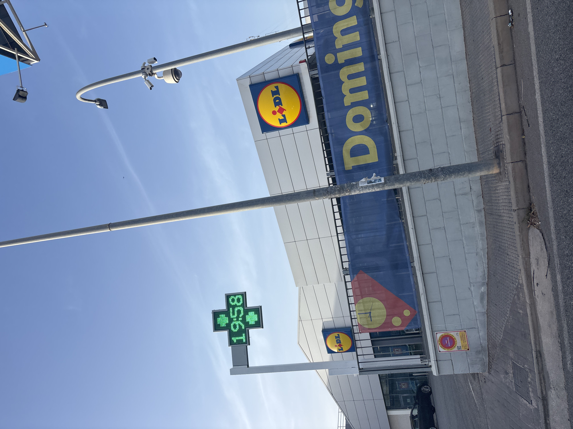 The image shows a street view in Orihuela, Spain, with a Lidl supermarket prominently featured. To the left of the Lidl building, a green digital cross sign displays the time "19:58". A blue banner with yellow lettering, partially obscured, reads "Domingu...". A black car is parked in front of the supermarket's entrance. In the foreground, a concrete sidewalk and asphalt road are visible, with a gray brick wall and a black metal fence separating the sidewalk from the supermarket's property. Overhead, a utility pole with a security camera and streetlights stands tall against a clear blue sky. Another utility pole is visible on the far right, with a glimpse of a blue overhang. A circular sign with a clock face is attached to the fence.