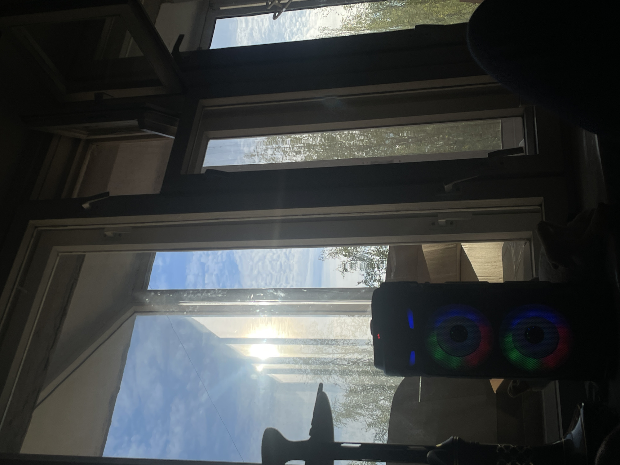 The image shows a view from inside a room looking out through a multi-paned window. The window reveals a daytime scene with a blue sky and clouds, along with trees visible in the distance. In the foreground, obscuring part of the view, is a dark, rectangular object with two circular illuminated areas, likely speakers. There is also an unidentifiable object in front of the window, towards the bottom of the frame. The scene appears to be indoors, with parts of the room's interior visible around the window frame. The location is unknown.