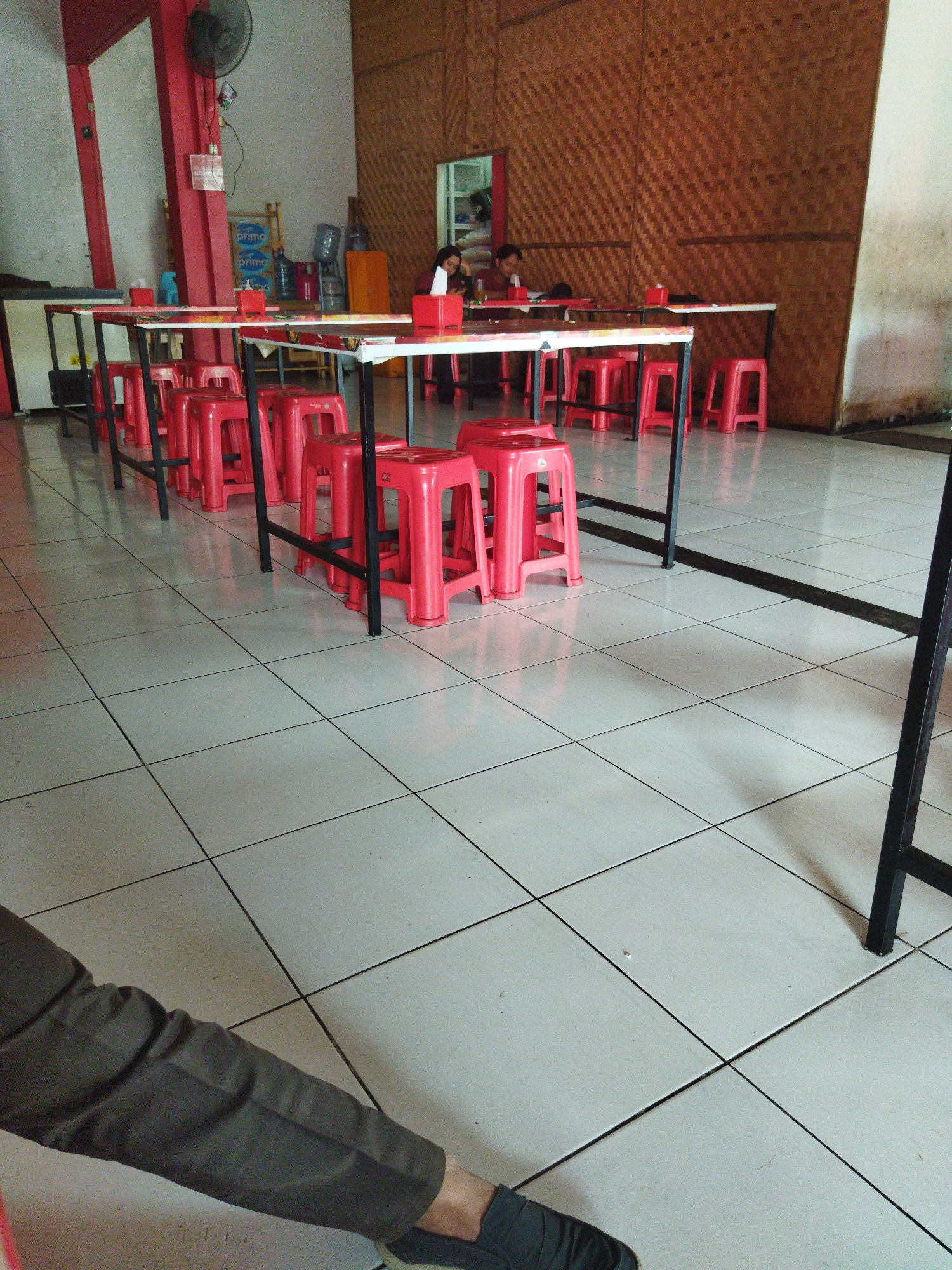 The image depicts the interior of a dining establishment, likely a warung or casual restaurant, with a focus on a row of tables and red plastic stools. The tables have white tops and black metal frames, and are set with red napkin holders. The floor is tiled in a light color with dark grout lines. In the background, a person is seated at one of the tables, seemingly eating or engaged in conversation. Another person is partially visible further down the row of tables. The walls are decorated with a woven rattan material. A ceiling fan is visible on the left side of the image. The overall impression is that of a simple, functional eatery. Based on the context of the image being in the city of Tangerang, Indonesia, this appears to be a typical local food stall or restaurant.