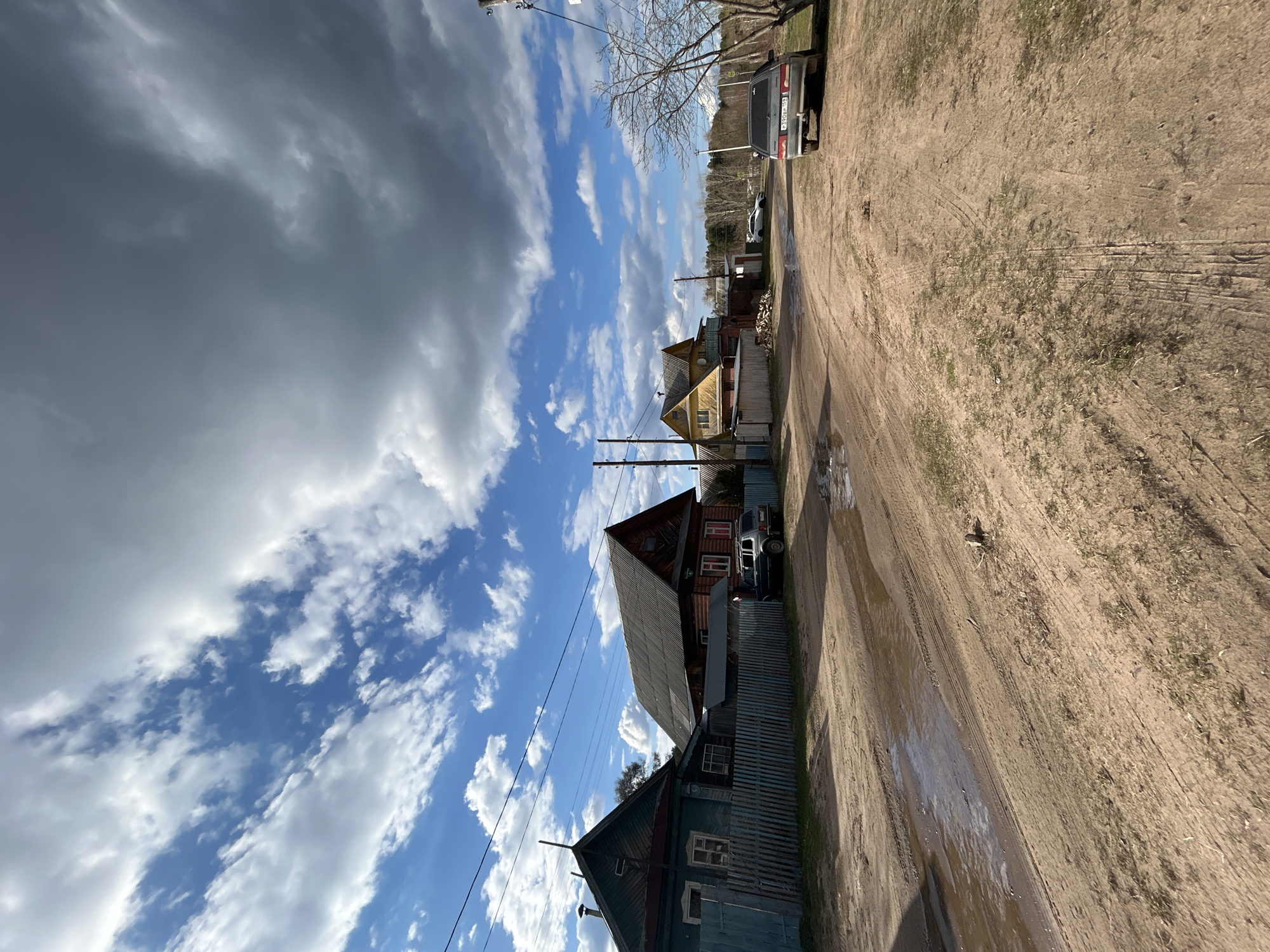 The image depicts a rural street scene in Chaikovsky, Russia. A dirt road runs along the right side of the frame, showing puddles and tire tracks. Several wooden houses of varying colors and conditions are lined up on the left, some with visible fences. Power lines stretch across the sky above the houses. A gray car is parked along the road, next to a house, as is another white car further in the background. The sky is filled with a mix of dark storm clouds and patches of blue sky with white clouds. Bare tree branches are visible in the upper part of the frame.