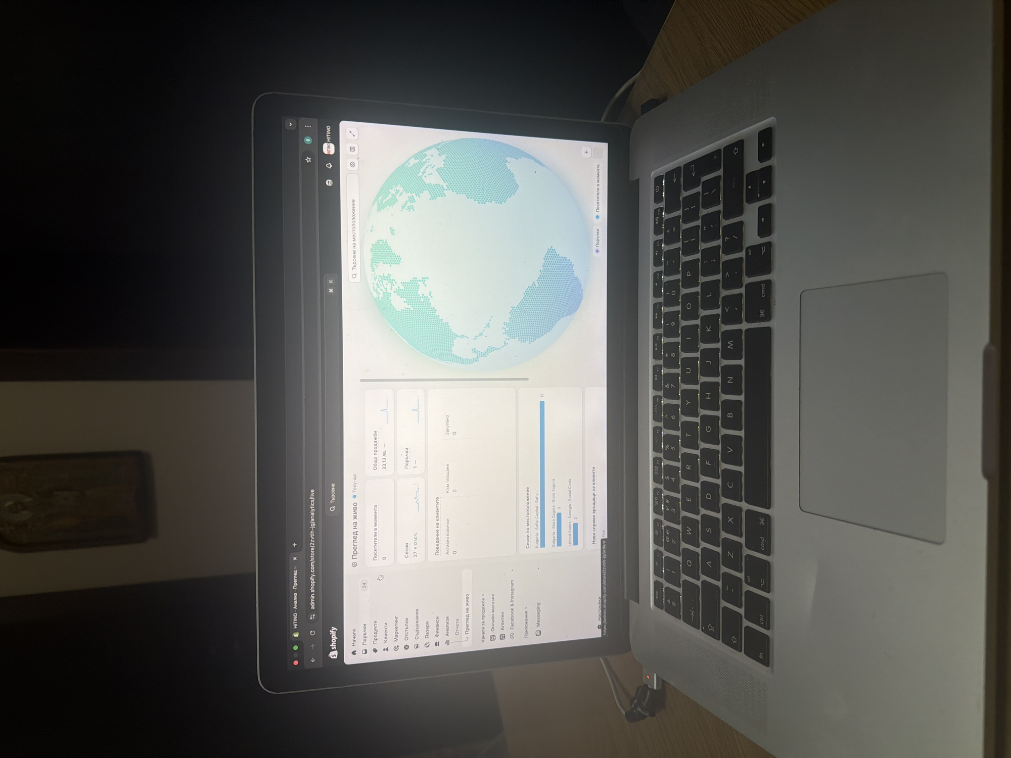 A silver MacBook laptop is captured from an elevated, angled perspective, resting on a light wooden surface in what appears to be an indoor setting in Shumen, Bulgaria. The background is dark and out of focus. The laptop's screen is open, displaying a Shopify e-commerce analytics dashboard, indicating an activity of managing or monitoring an online store.
The screen's main feature is a world map visualization, depicted with turquoise and blue hexagons, likely illustrating global website visitor locations. Below the map, various metrics are presented in Bulgarian. Key data points visible include "Общо продажби" (Total sales) at "33,13 лв." (33.13 BGN), "Поръчки" (Orders) at 1, and "Сесии" (Sessions) at 27. The section for "Посетители в момента" (Visitors at the moment) shows 0.
Under "Сесии по местоположение" (Sessions by location), specific geographic data is displayed: "Bulgaria Sofia-Capital Sofia" (3 sessions), "Bulgaria Stara Zagora Stara Zagora" (2 sessions), and "United States Georgia Social Circle" (1 session). The browser's URL bar shows `admin.shopify.com/store/2zvtih-jg/analytics/live`, confirming the platform. The left sidebar of the dashboard lists various Shopify administrative sections such as "Начало" (Home), "Поръчки" (Orders), "Продукти" (Products), "Анализи" (Analytics), and "Преглед на живо" (Live view). The overall appearance suggests active management of an e-commerce business.