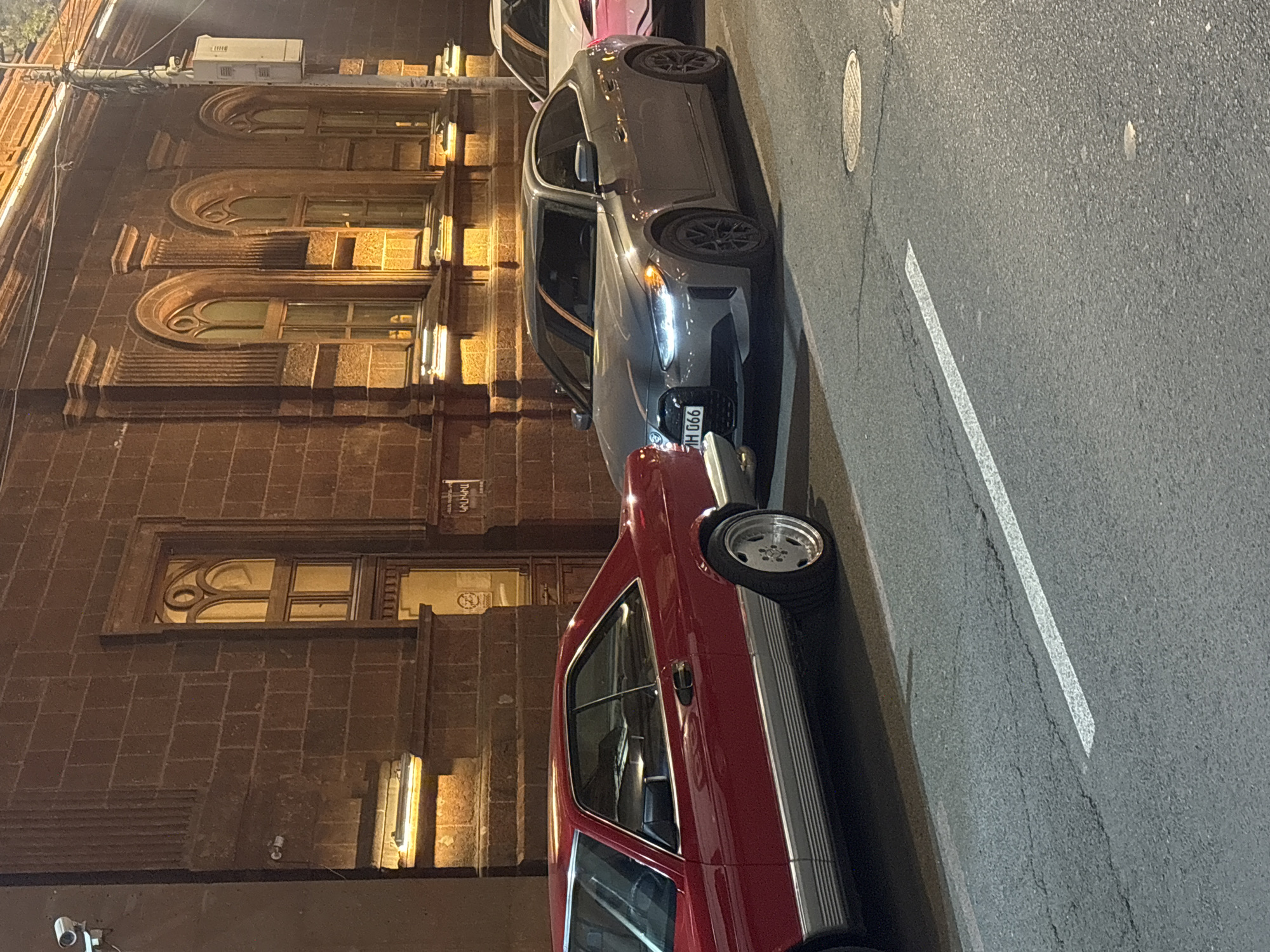 The image depicts a nighttime street scene in Yerevan, Armenia. The photograph is taken from a slightly elevated perspective, facing a building with ornate window designs and a brick facade. Two parked cars are visible alongside the curb: a red classic car with chrome accents closest to the viewer and a grey modern sedan with license plate number "990 H" parked behind it. The street is paved asphalt with a white lane marking, and a manhole cover is visible in the foreground. Street lighting is provided by the building's facade lights. No people are visible in the image.