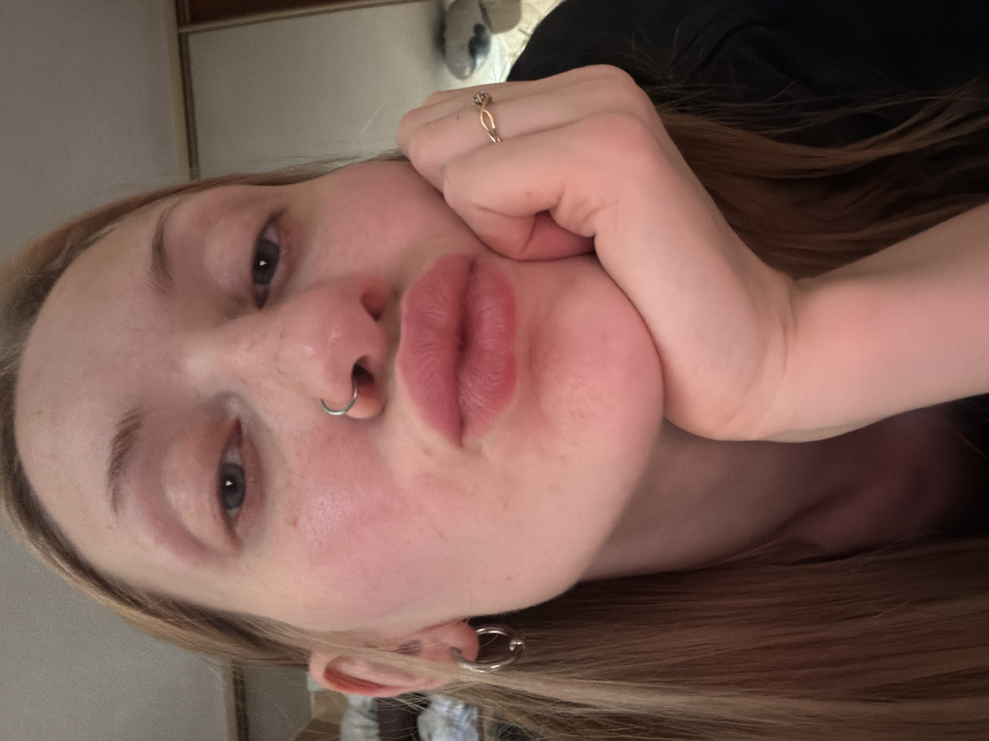 A close-up, slightly angled, selfie-style photo shows a young woman with fair skin, blonde hair, and a pinkish complexion. She is wearing a nose ring and a hoop earring in her left ear. Her right hand, adorned with a gold ring with a small stone, is resting on her chin, with her thumb tucked into her palm. Her lips are pursed in a kissing gesture, and her eyes are open, looking directly at the camera. The background is blurred, but suggests an indoor environment with a neutral-colored wall and possibly furniture. There are no discernible elements that would place this image in Saint Petersburg, Russia.