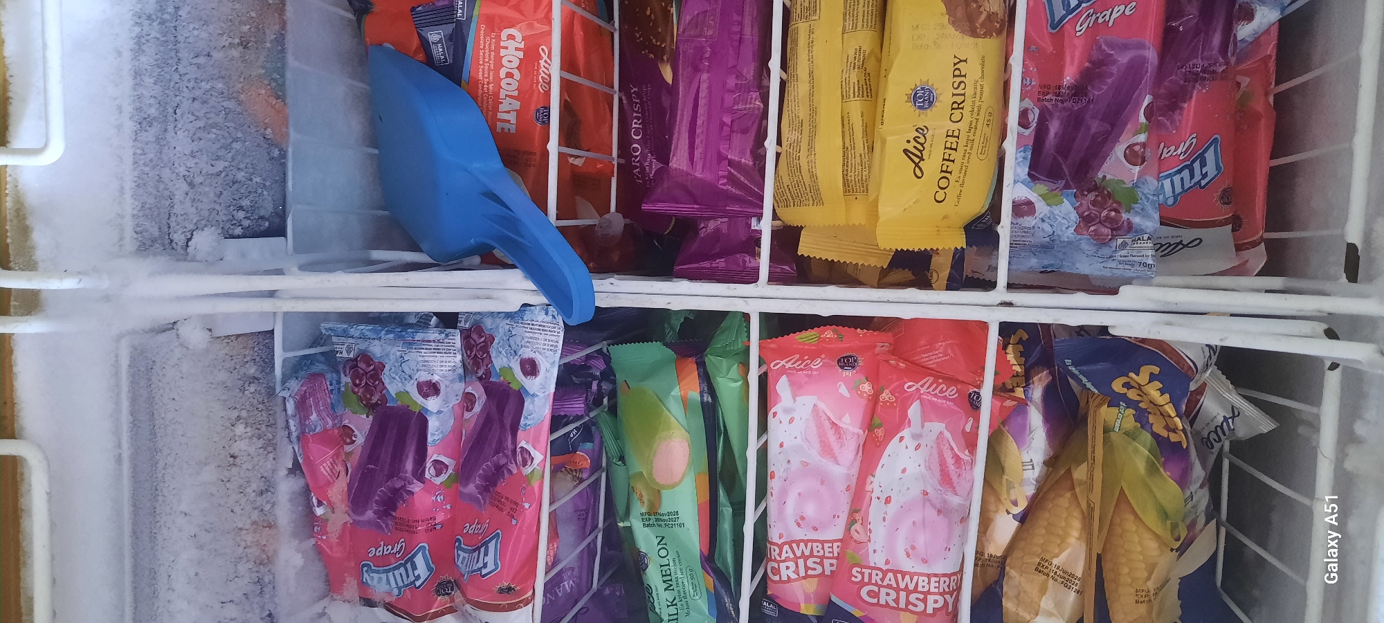 This is a photograph taken inside a freezer, likely in the city of Selopuro, Indonesia. The freezer is filled with various ice cream bars and other frozen treats, predominantly packaged in brightly colored wrappers. Some of the visible flavors include grape, milk melon, strawberry crispy, and coffee crispy. There are also some less clearly identifiable items that appear to be corn-flavored ice cream.
A bright blue plastic ice cream scoop is resting on top of some of the packages, suggesting recent use or preparation for serving. The interior of the freezer shows signs of frost accumulation on the walls and the metal shelving, indicating a typical frozen food storage environment. The lighting is likely artificial, consistent with indoor lighting. The image is a close-up, focusing on the contents of the freezer, with a portion of the freezer door or frame visible at the top and bottom edges. The "Galaxy A51" watermark at the bottom left indicates the device used to capture the image.